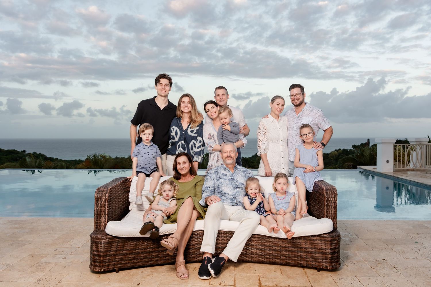 Group gathered on wicker outdoor furniture by an infinity pool at sunset with cloudy sky in background.