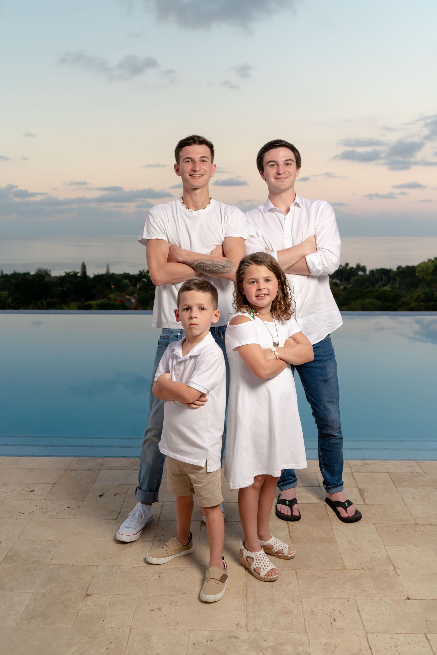 Four family members pose together in white outfits by an infinity pool at sunset against a cloudy sky.