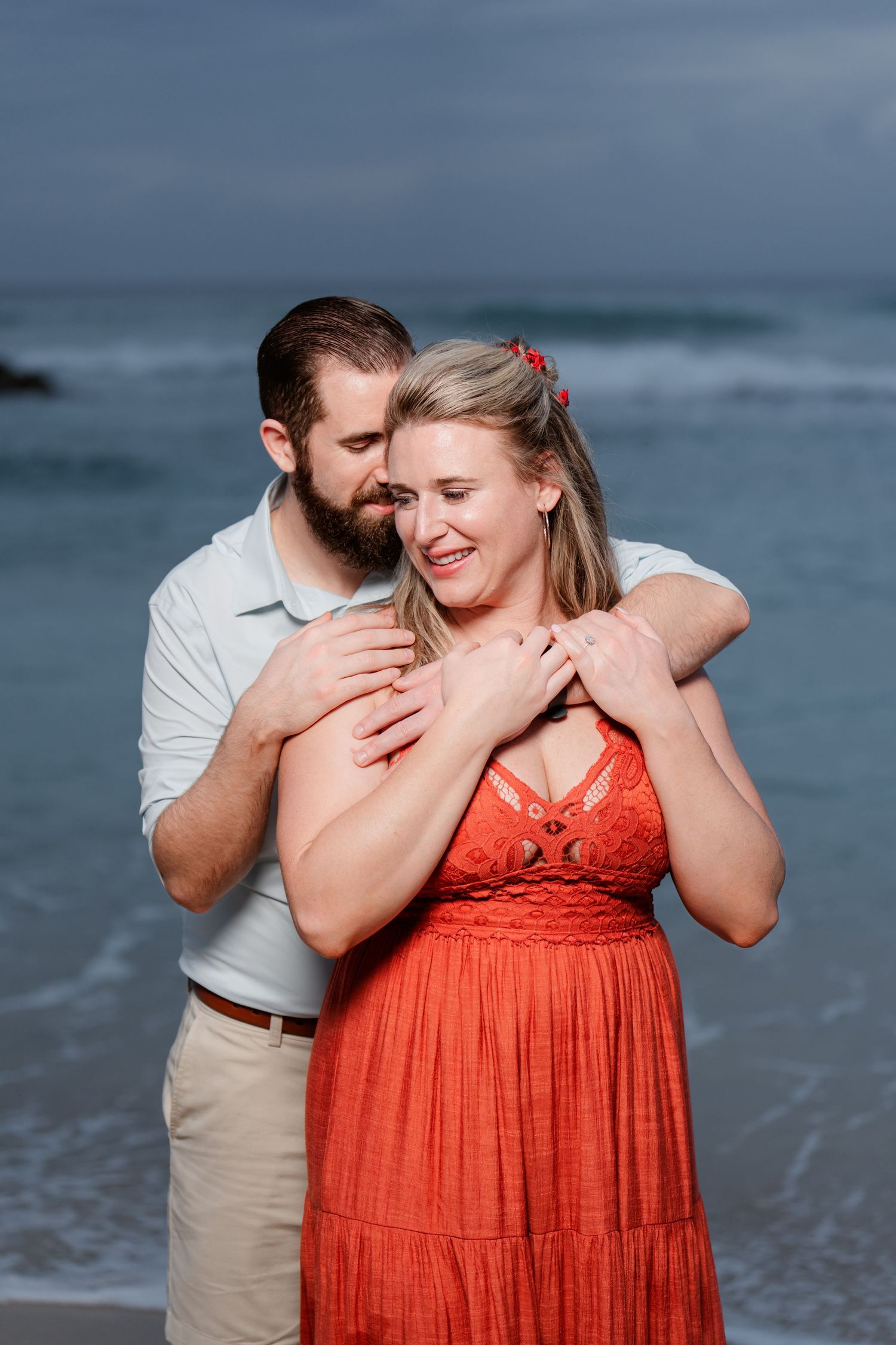 A romantic couple embraces on a cloudy beach, with her wearing a coral dress and him in a white shirt and khakis.