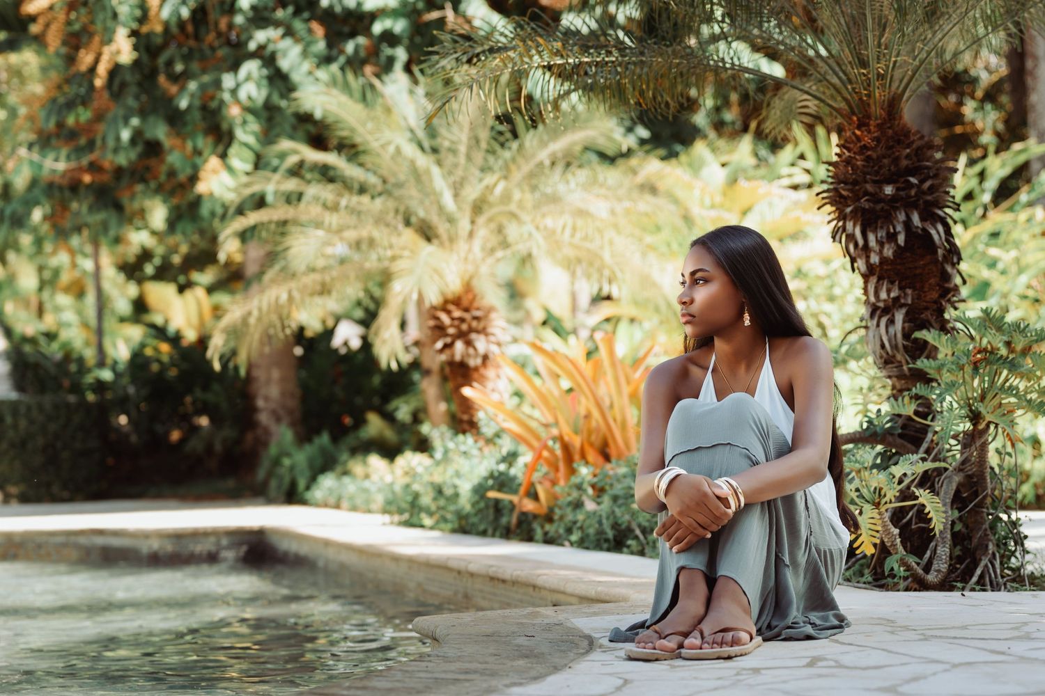A couple sits together on stone steps surrounded by lush tropical plants and palm fronds in a garden setting.