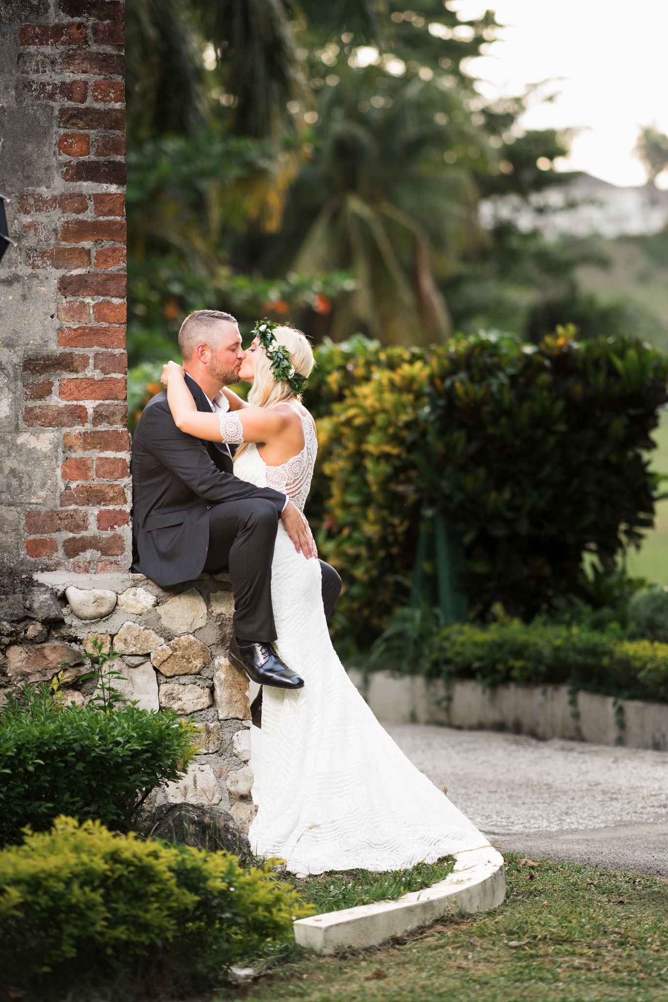 Bride and groom share a romantic kiss near a rustic brick wall surrounded by lush tropical greenery.