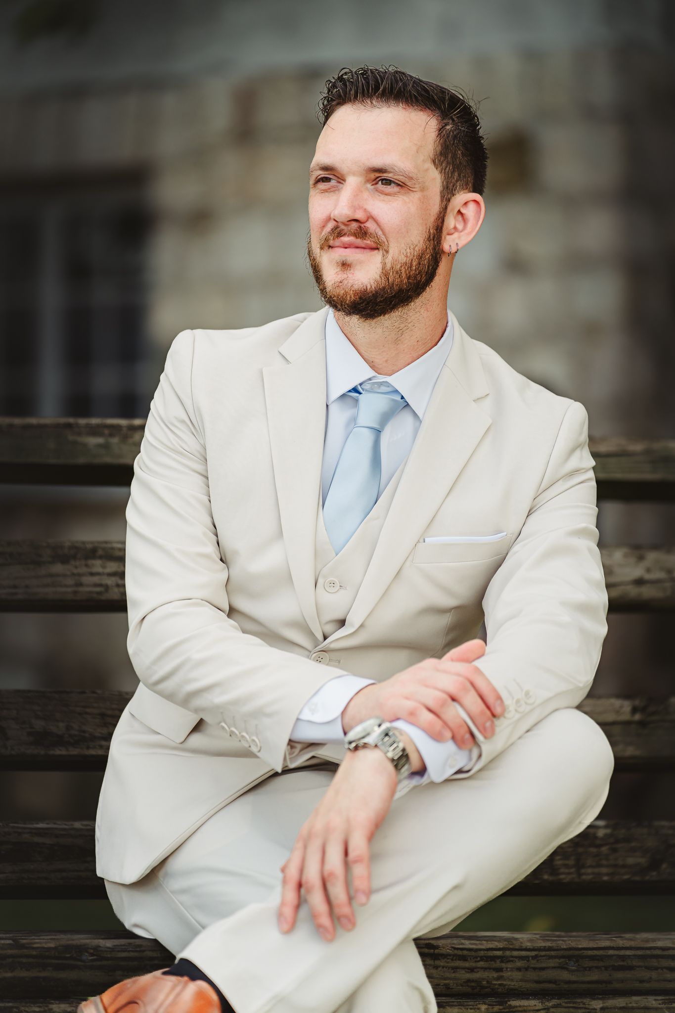Smiling bearded man in beige suit and light blue tie sitting on wooden bench outdoors.
