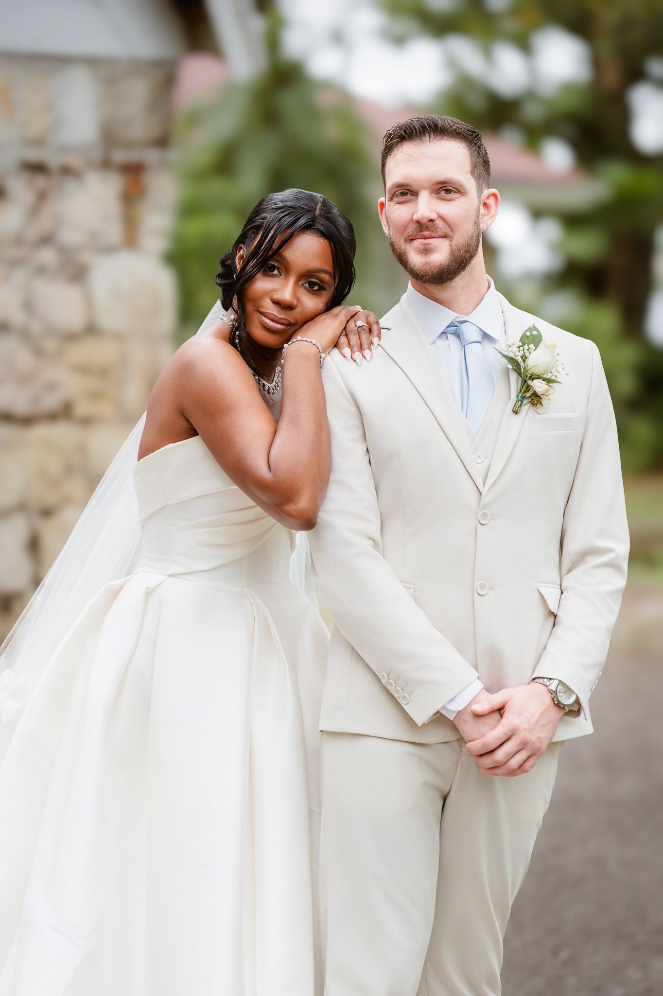 Interracial couple poses outdoors on wedding day, bride in strapless white gown leans on groom in cream suit.