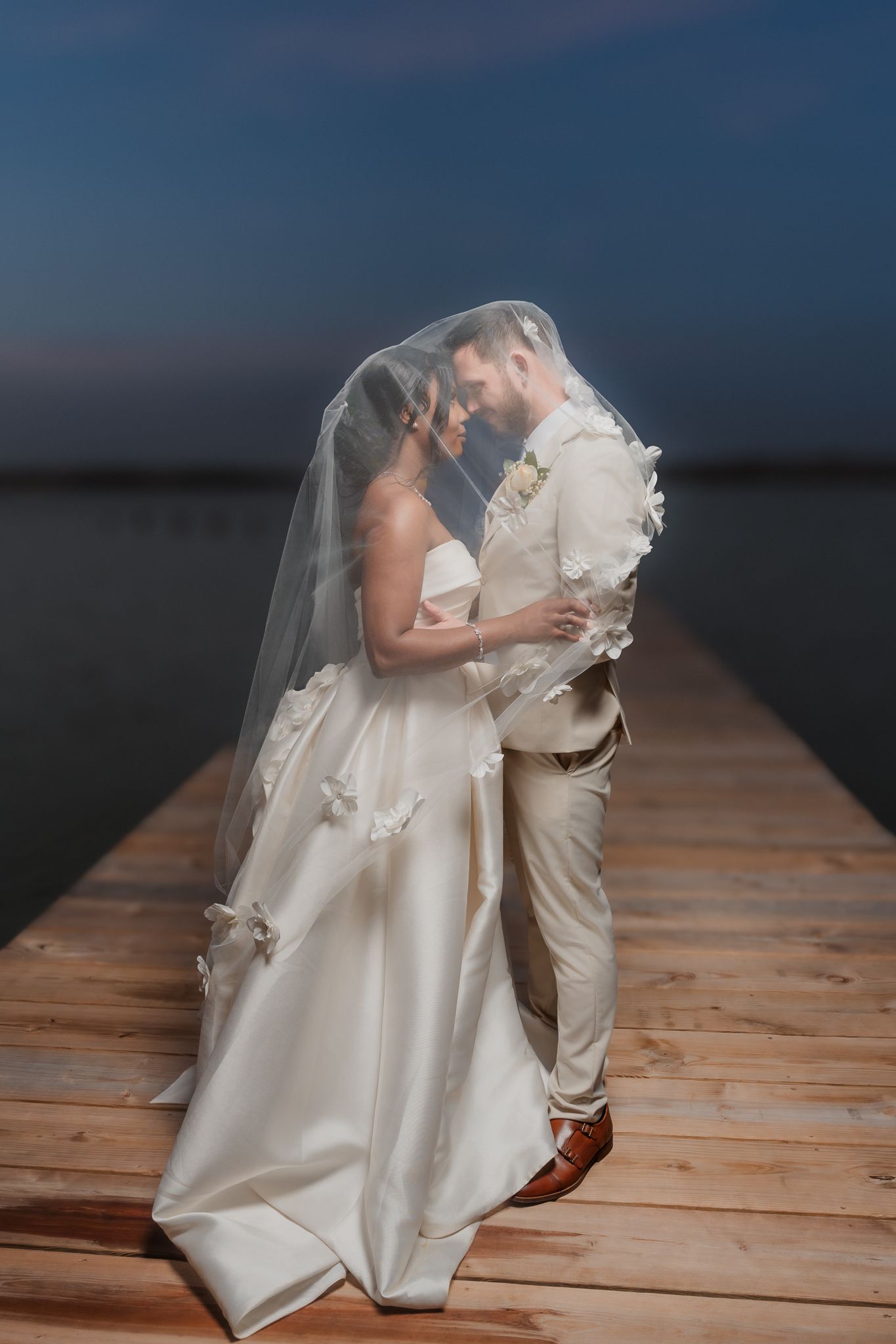 Bride and groom share romantic moment under veil on wooden dock at dusk, with calm water in background.