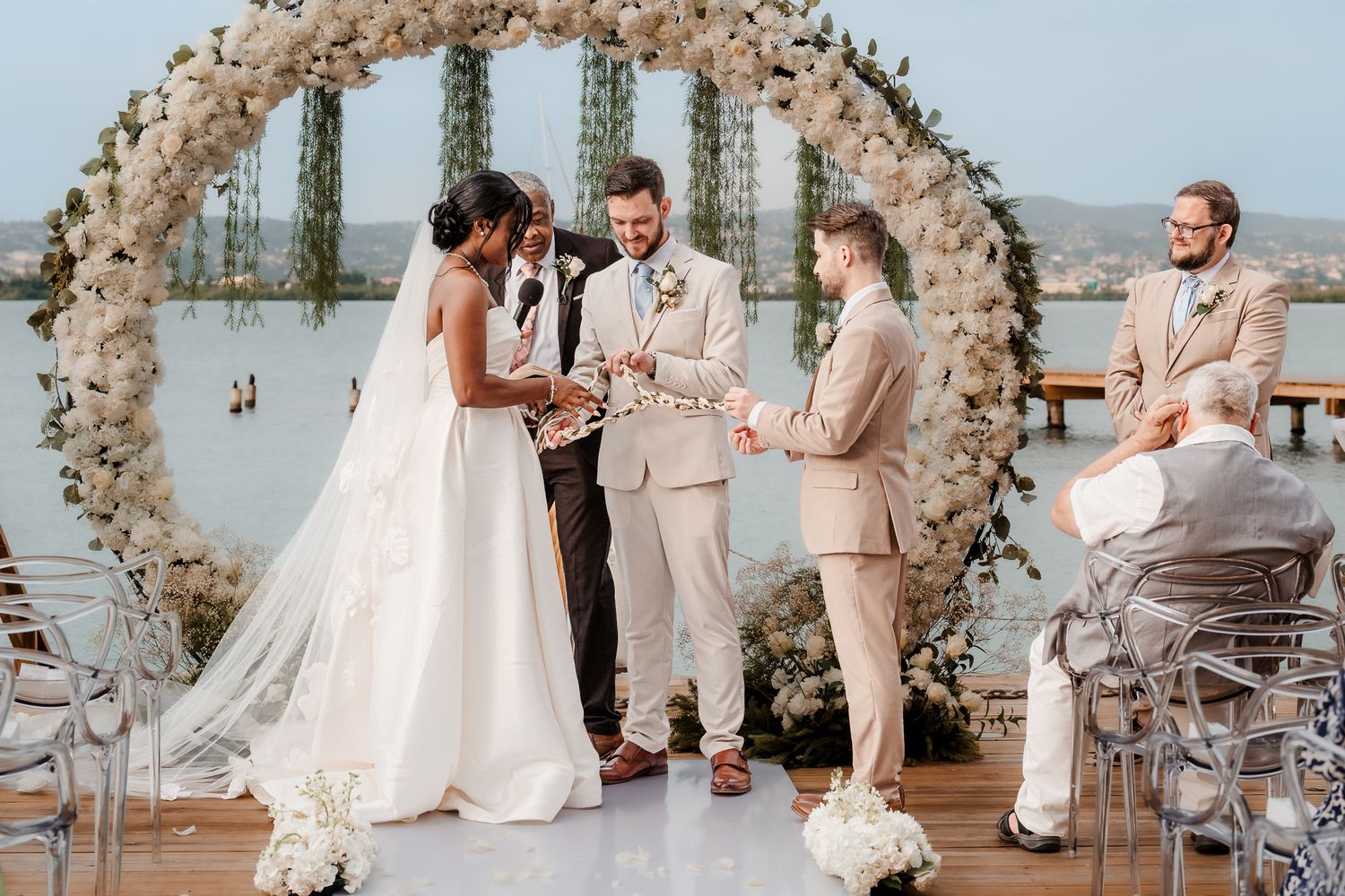 Outdoor wedding ceremony with bride and groom exchanging vows under a floral arch by the water.