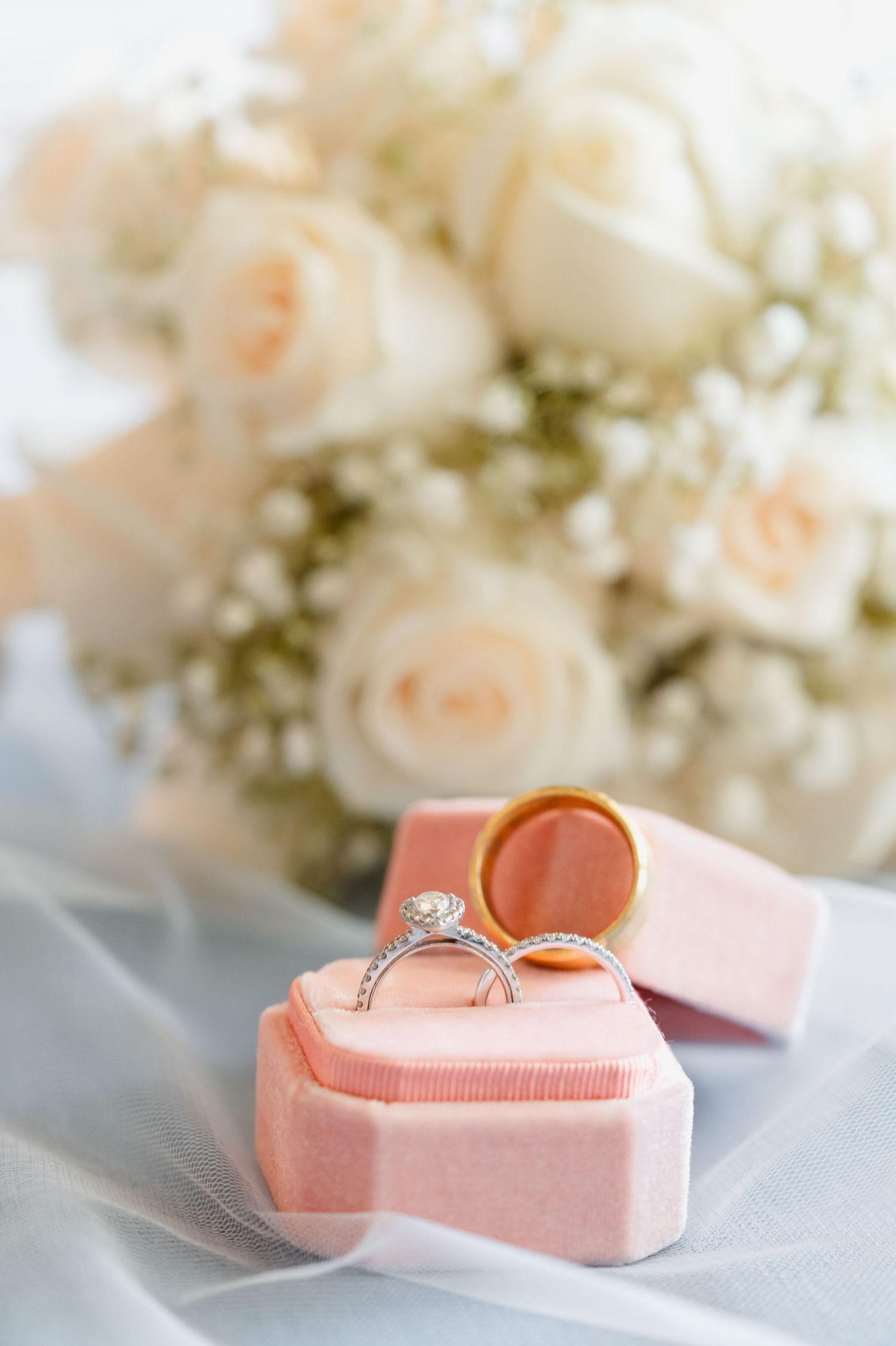 Wedding rings rest in a pink heart-shaped box with delicate cream roses and baby's breath flowers in soft focus background.