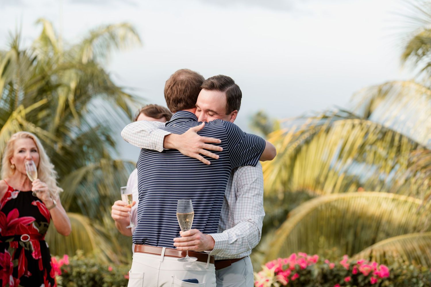 People embrace at an outdoor celebration with palm trees and tropical flowers in the background while holding champagne glasses.
