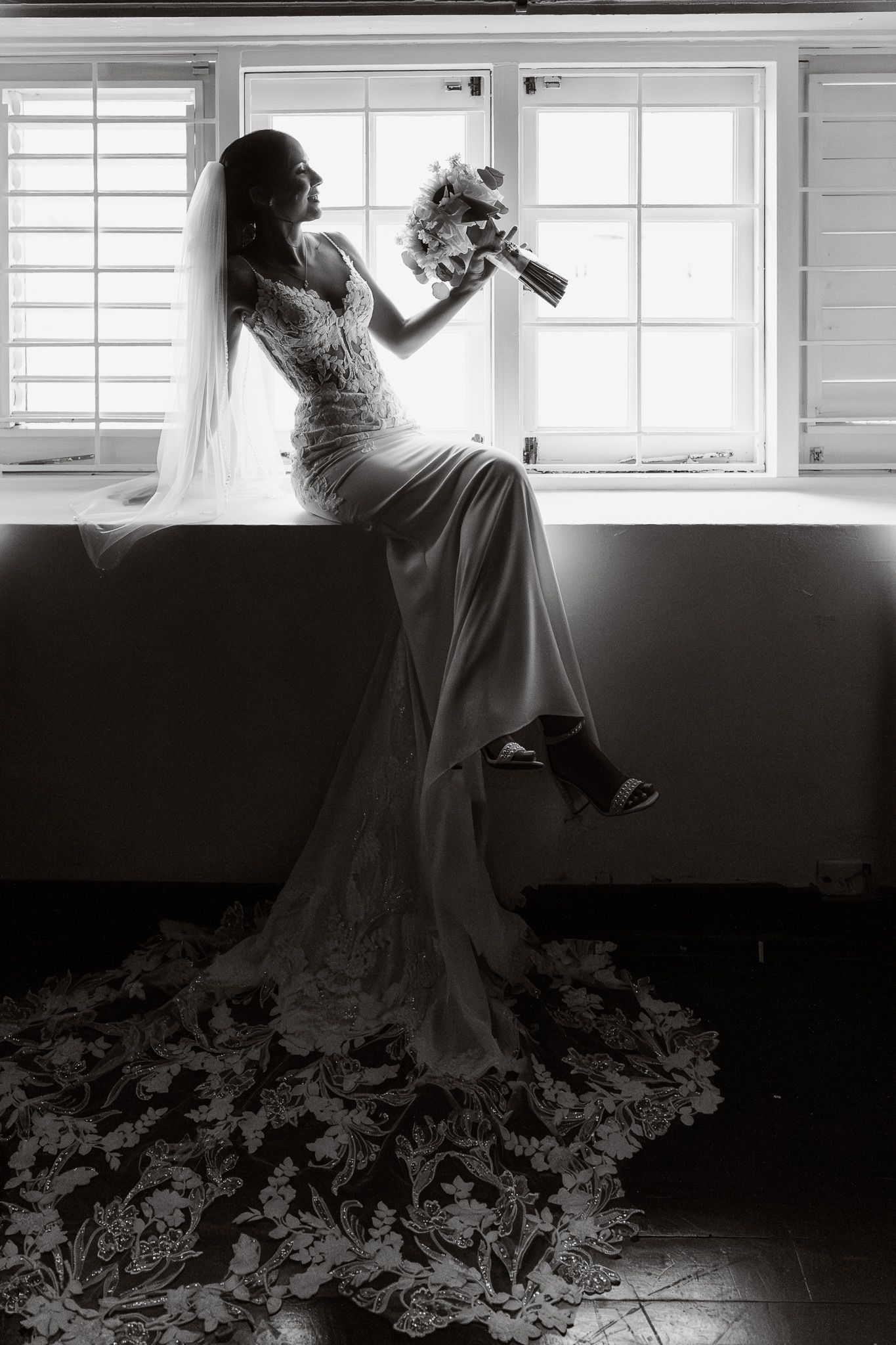 Black and white series of a bride in silhouette drinking champagne near a window with dramatic lighting.