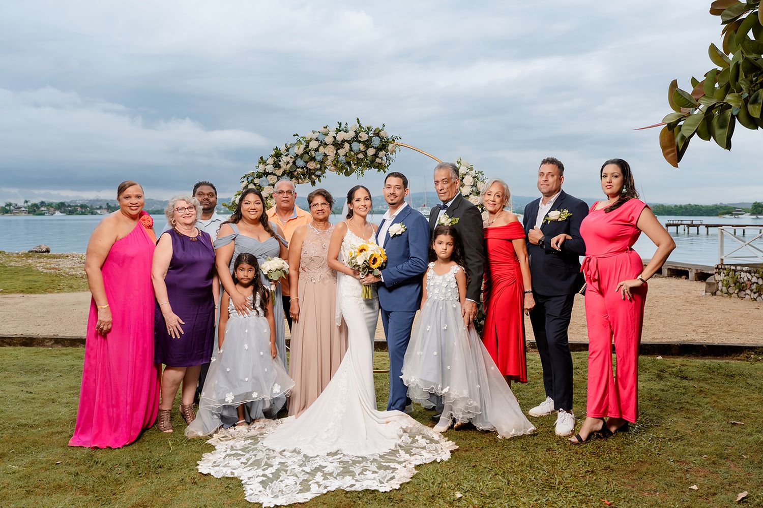A wedding party in colorful formal attire poses together outdoors on a waterfront lawn under cloudy skies.