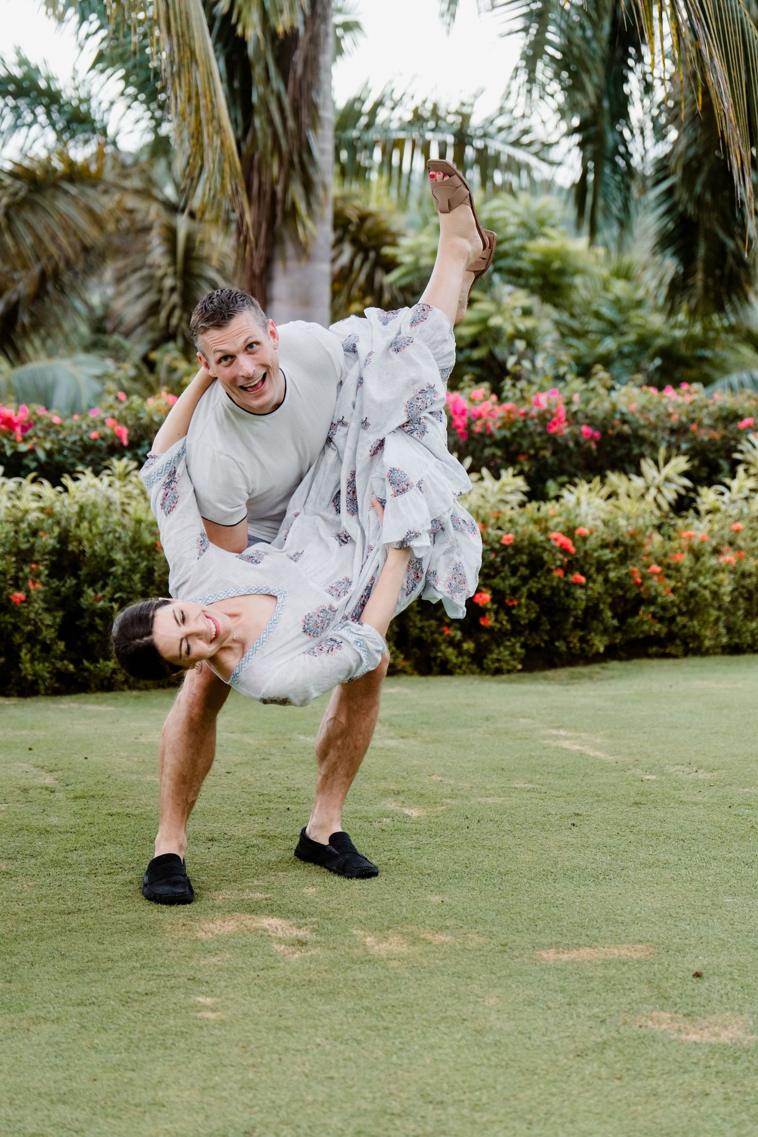 A couple wearing matching floral outfits dances playfully on a green lawn with tropical palm trees and flowers in background.