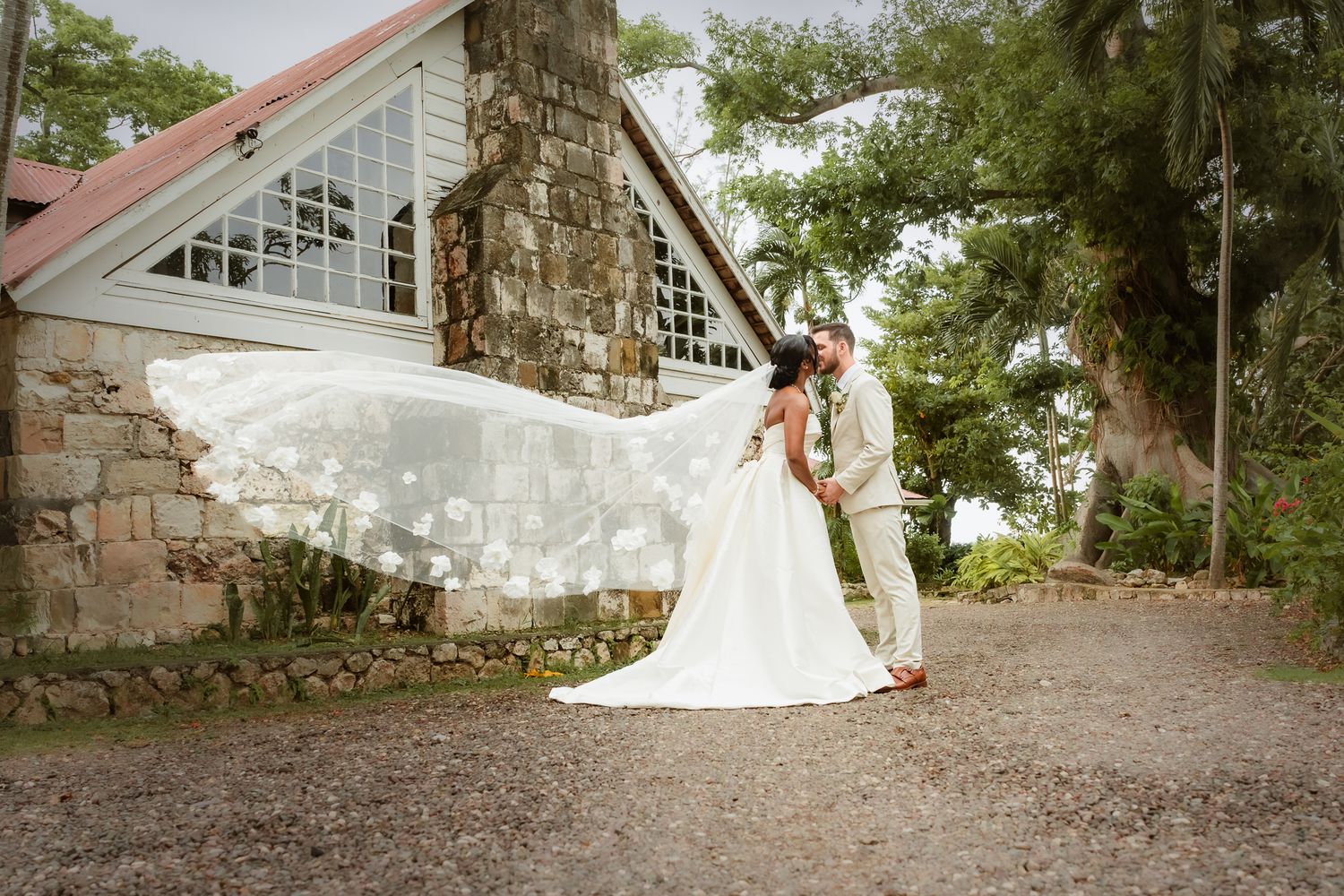Bride and groom share a romantic kiss outdoors, with veil flowing in the wind beside a stone chapel.