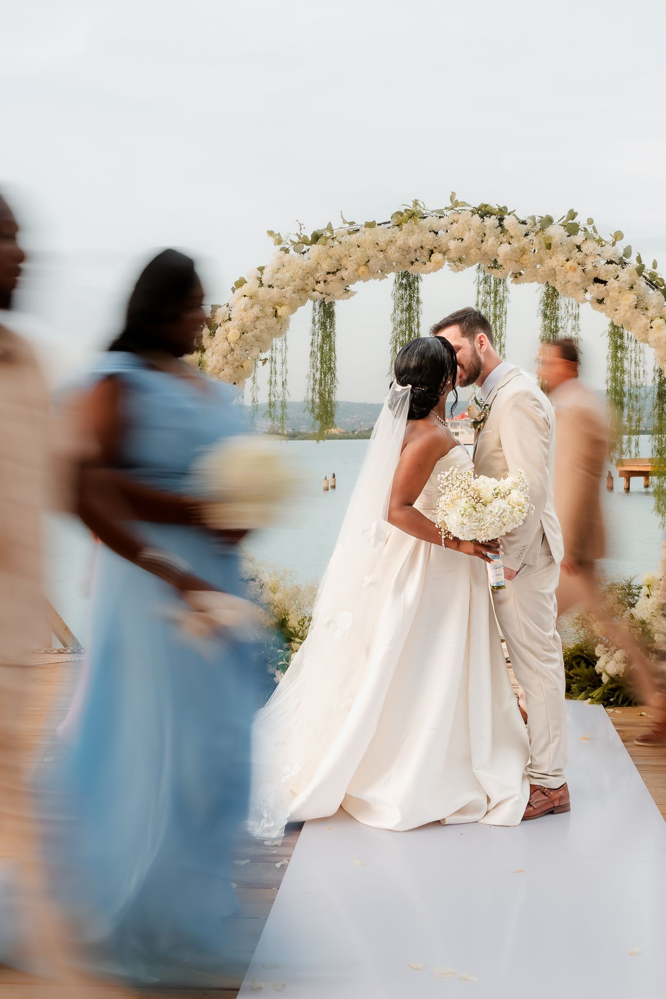 Bride and groom share a kiss at outdoor wedding ceremony with white floral arch, as bridesmaids in blue dresses walk by.