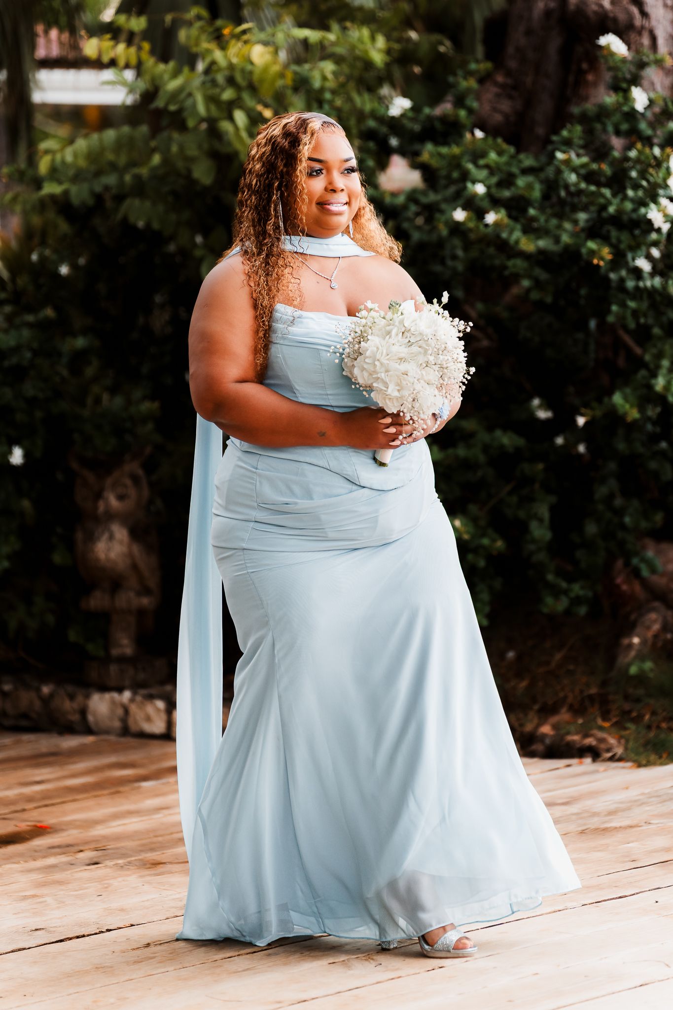 Plus-size bridesmaid in light blue chiffon gown holding white floral bouquet, smiling outdoors at wedding.