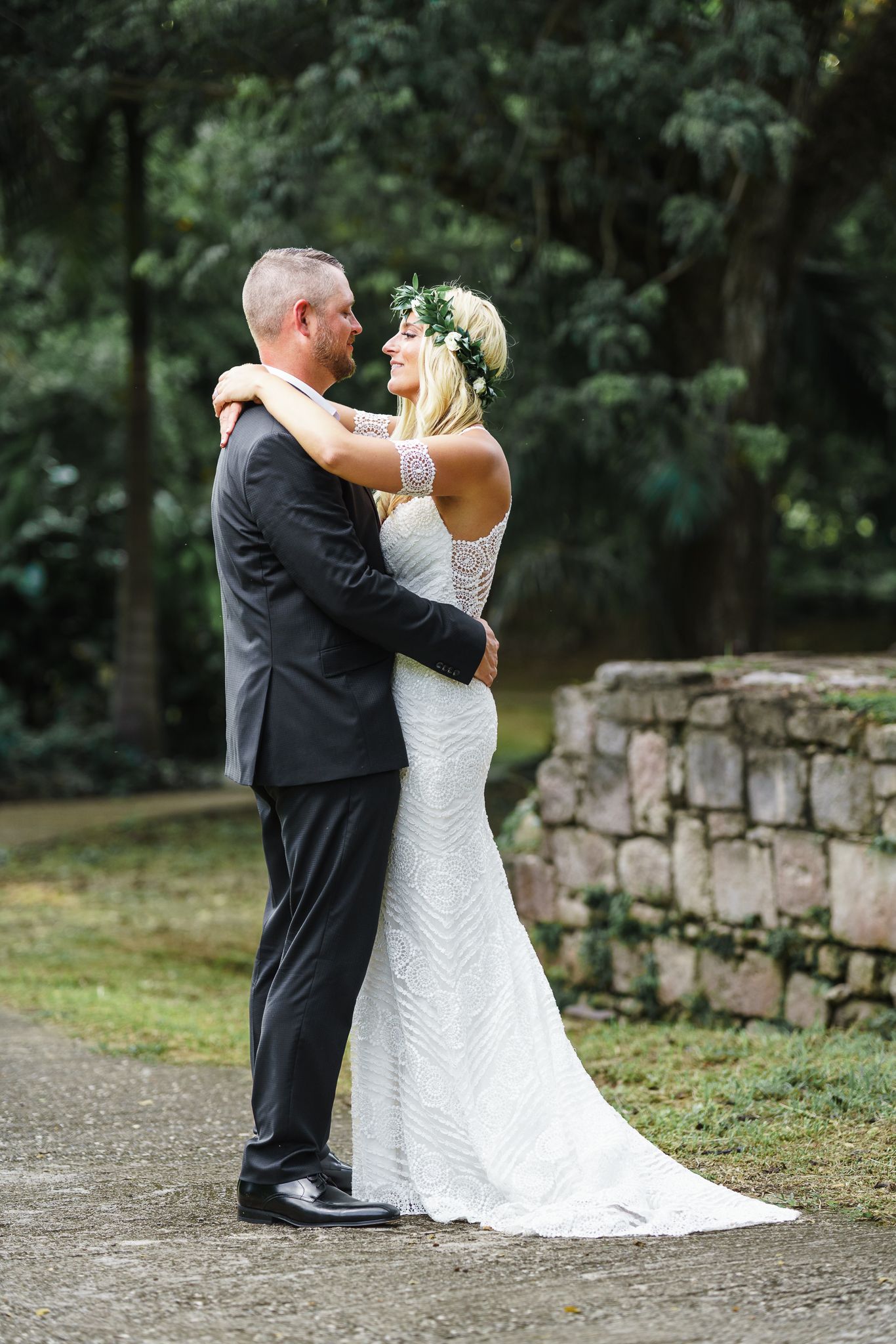 A bride in a white lace gown and floral crown embraces her groom in a black suit outdoors near stone wall.
