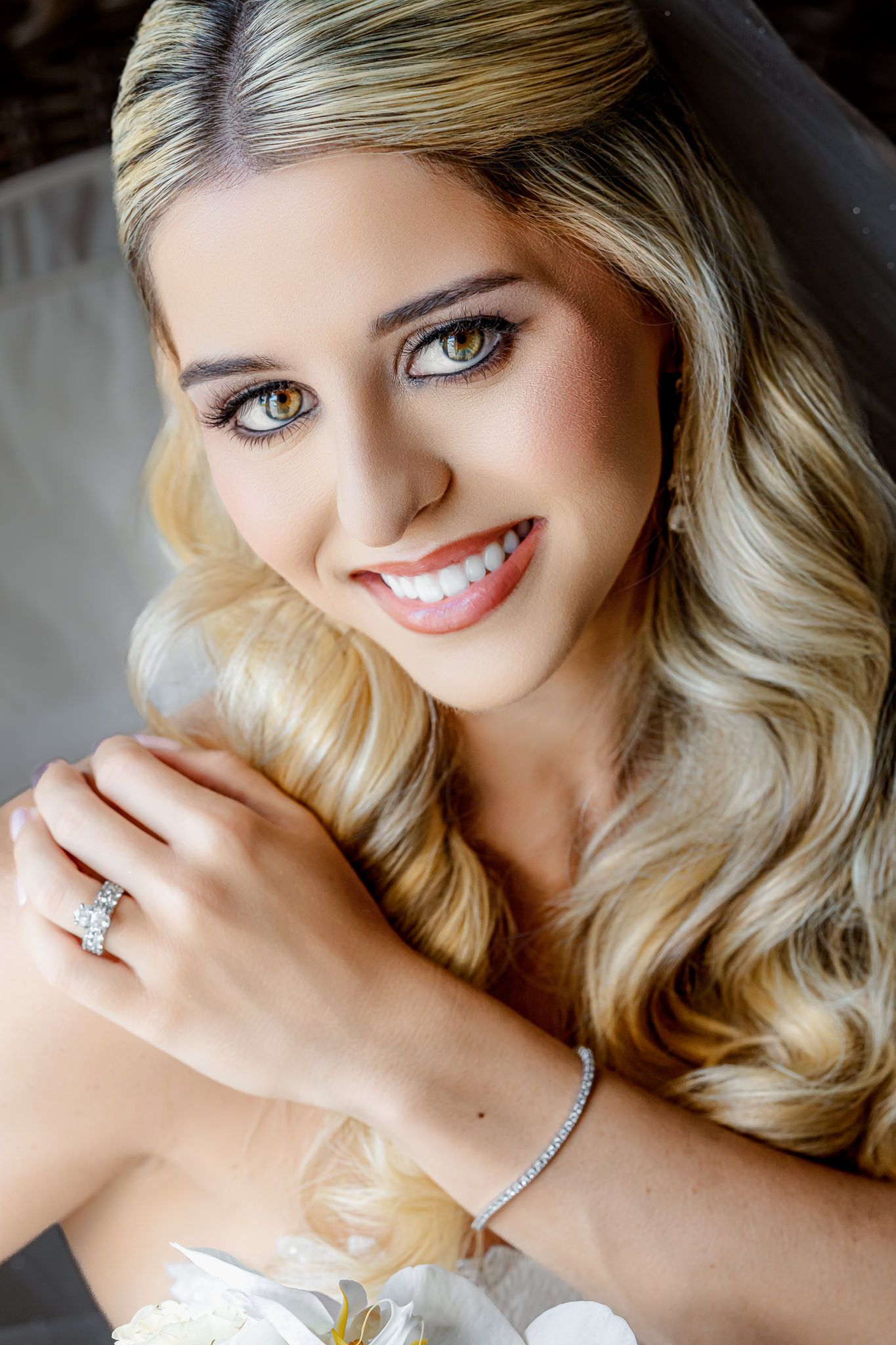 Smiling blonde bride with wavy hair, diamond ring and bracelet, wearing a white wedding dress in a elegant bridal portrait.