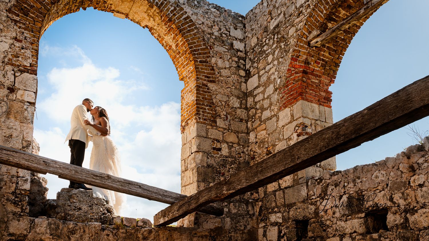 Bride and groom kissing in ancient stone ruins with arched walls and wooden beams under a blue sky.