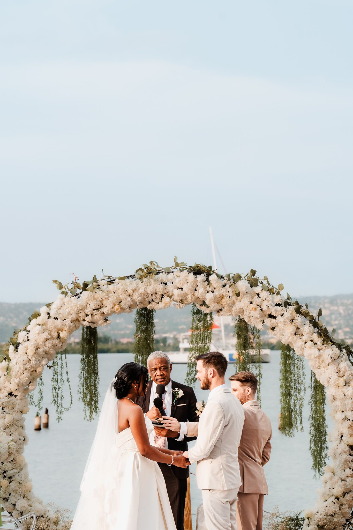 Couple exchanges vows under floral arch with officiant, waterfront backdrop, outdoor wedding ceremony.