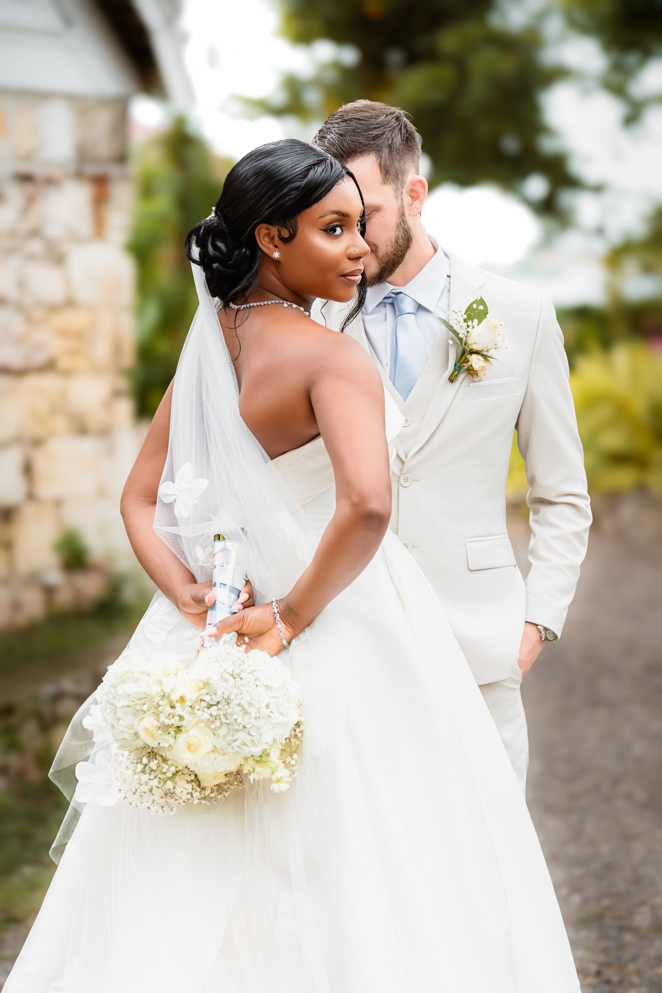 Interracial couple in wedding attire, bride in white gown holding bouquet, groom in cream suit, sharing intimate moment outdoors.