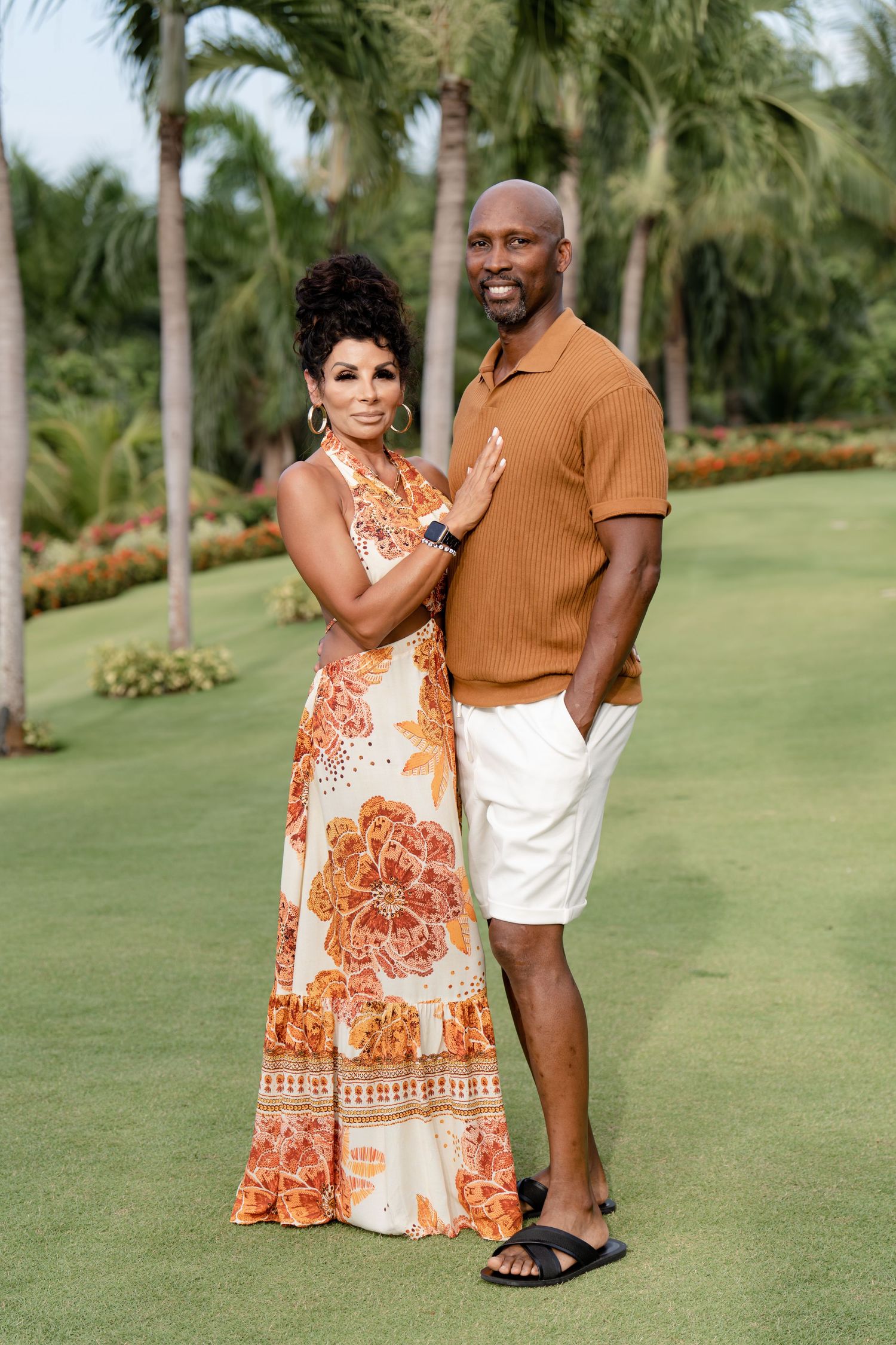 A couple poses on a lush golf course with palm trees, wearing coordinated orange and white resort attire.
