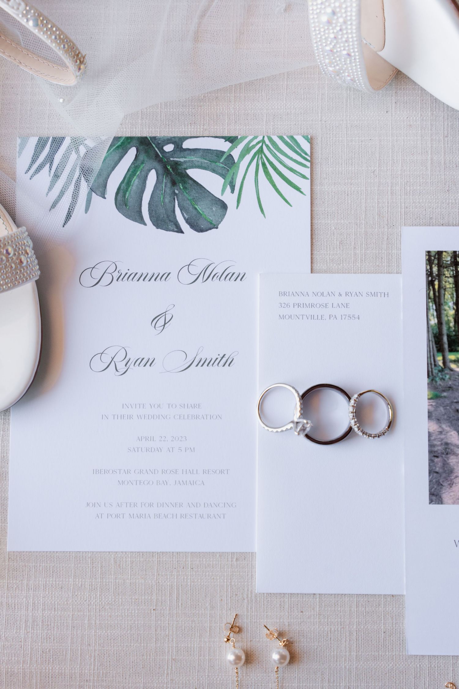 Wedding invitation with tropical leaf design next to wedding rings and pearl earrings on beige linen fabric.