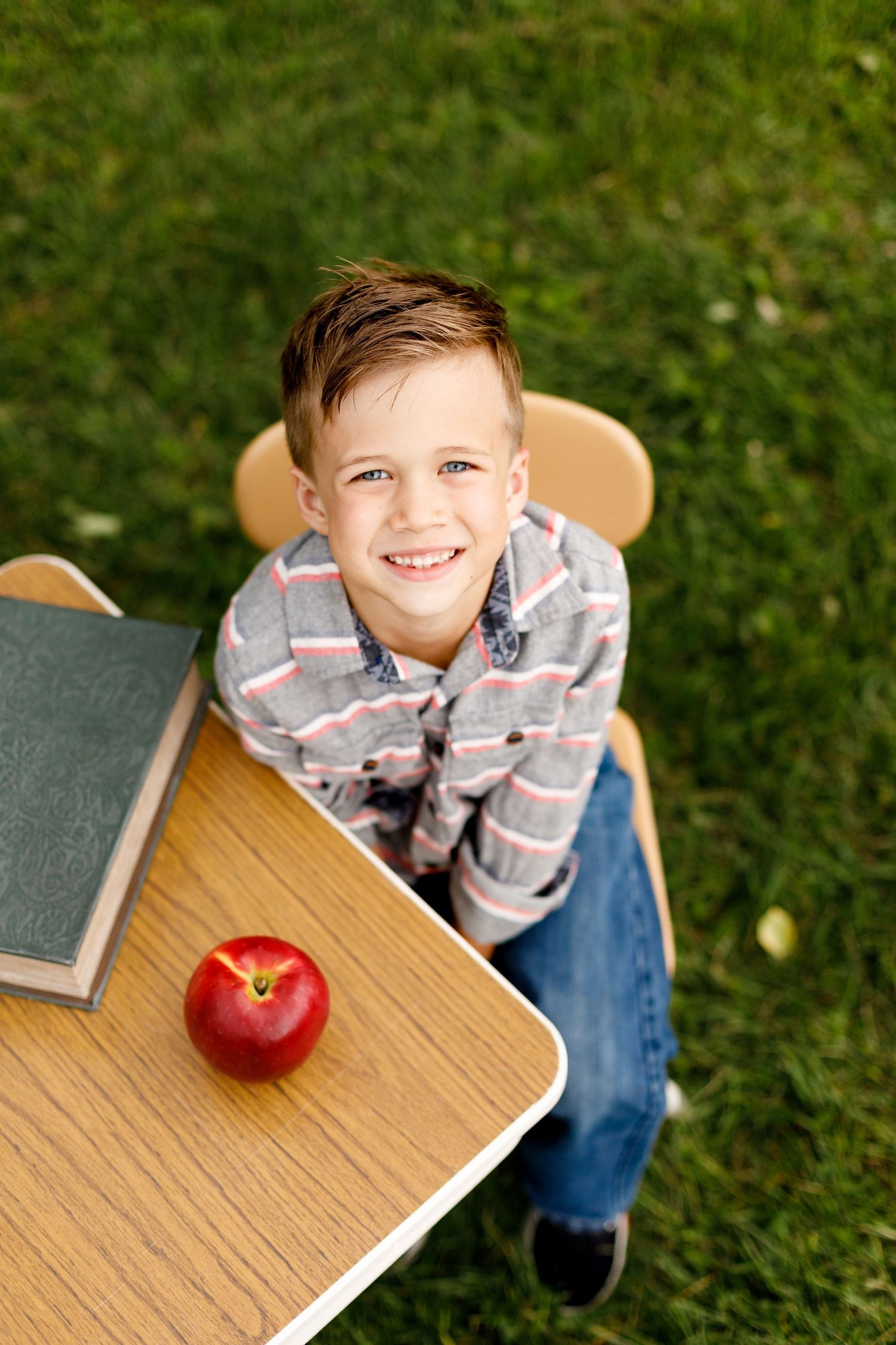 Books and a fresh red apple sit on a wooden desk in an outdoor setting with green grass.