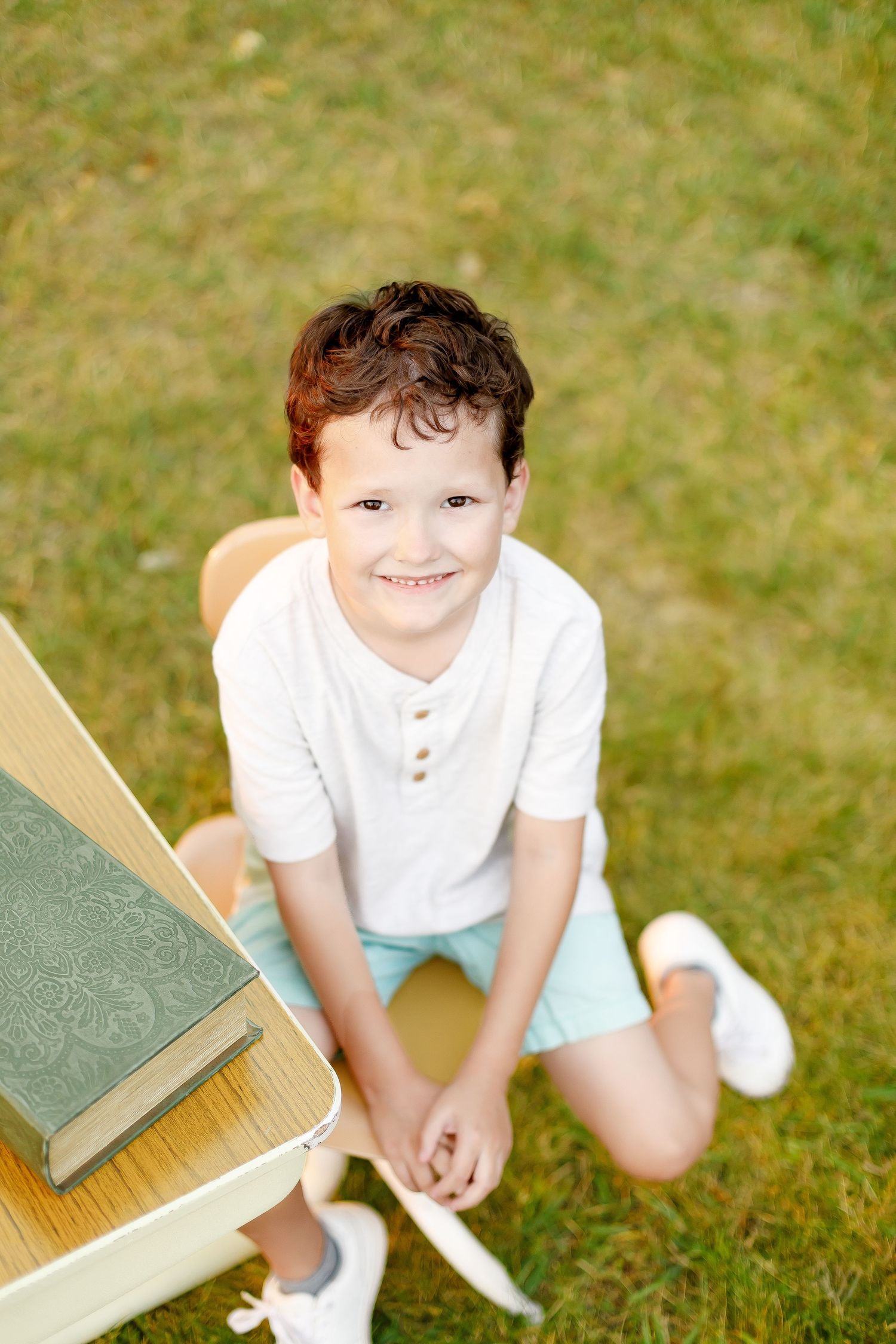 Child in white polo shirt posing playfully with vintage school desk outdoors.