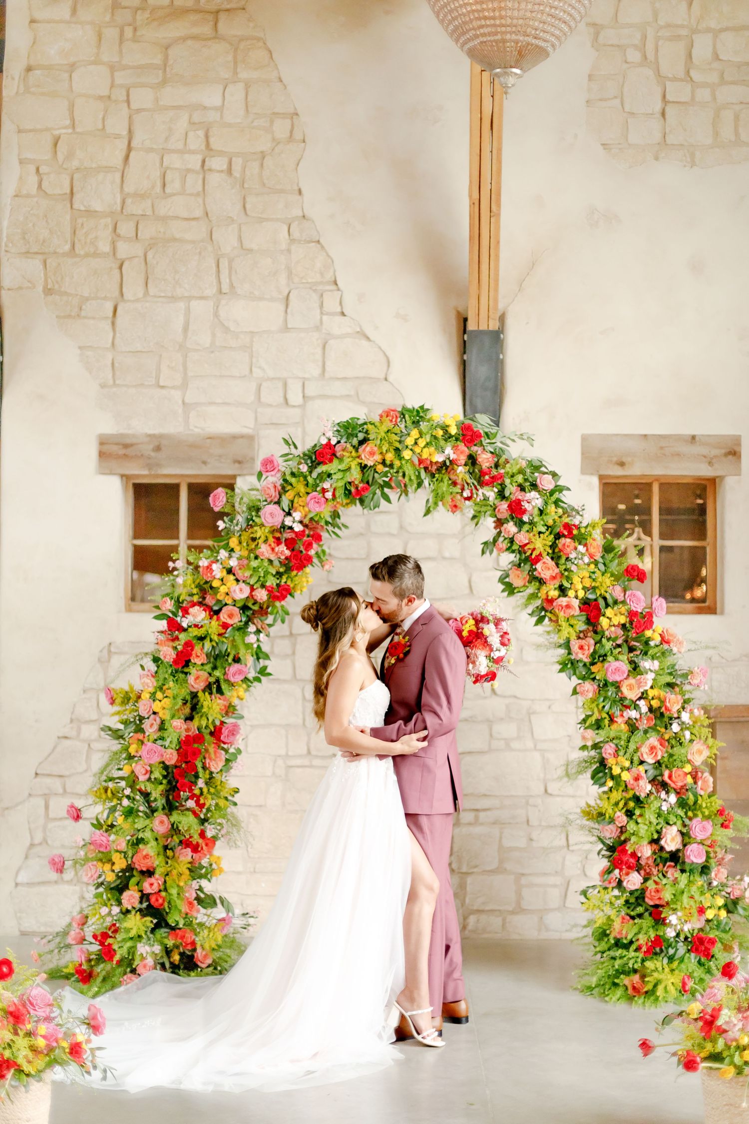 Couple embraces under beautiful floral arch with pink and red flowers against white stone wall at indoor venue.