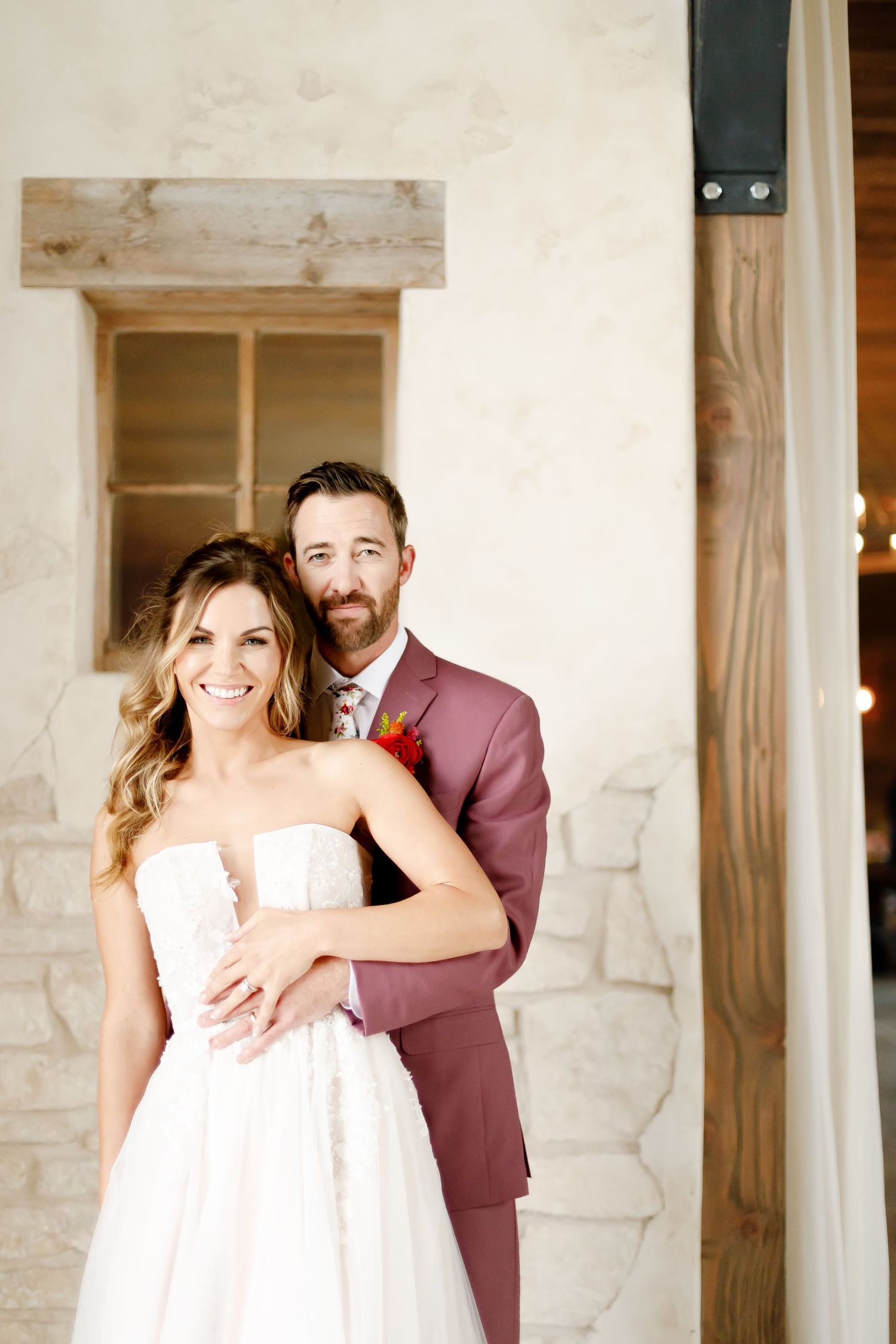 A wedding couple embraces on a rustic porch, with the bride in white and groom in burgundy suit.