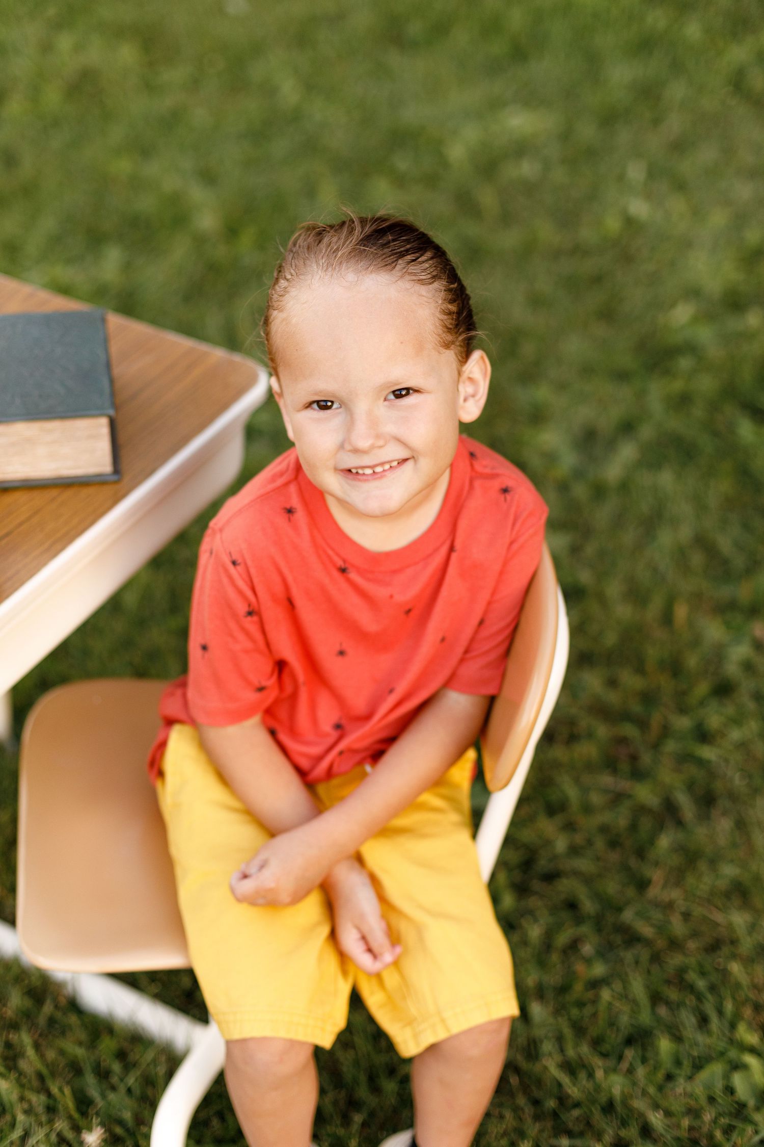 A young child in a bright red shirt and yellow shorts sits on light-colored furniture outdoors.