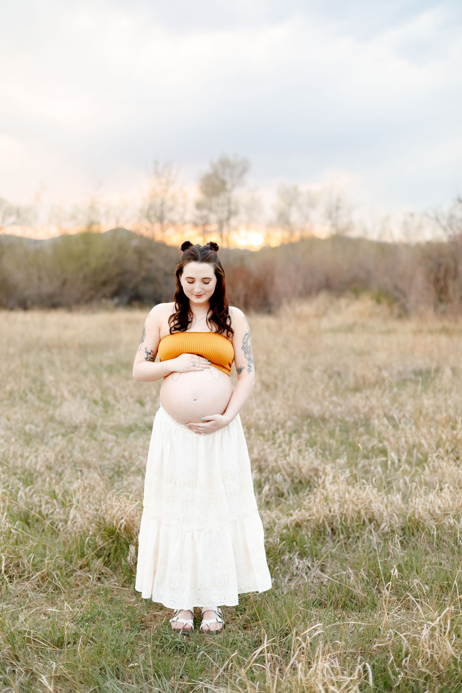 A maternity photo in a grassy field at sunset shows an expectant mother in a mustard top and white maxi skirt.