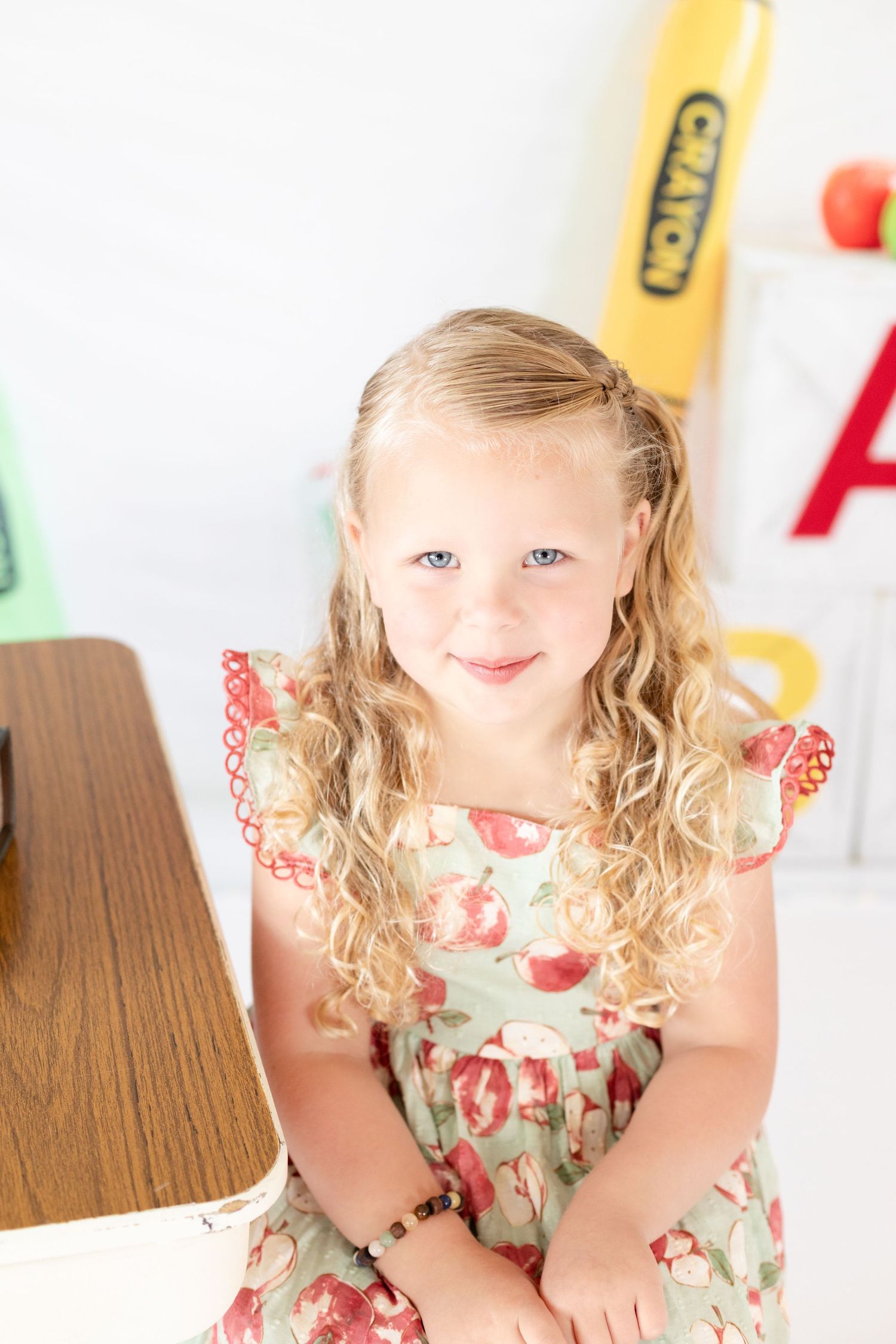 Young child wearing a floral dress sits at a wooden table with colorful decorations in the background.