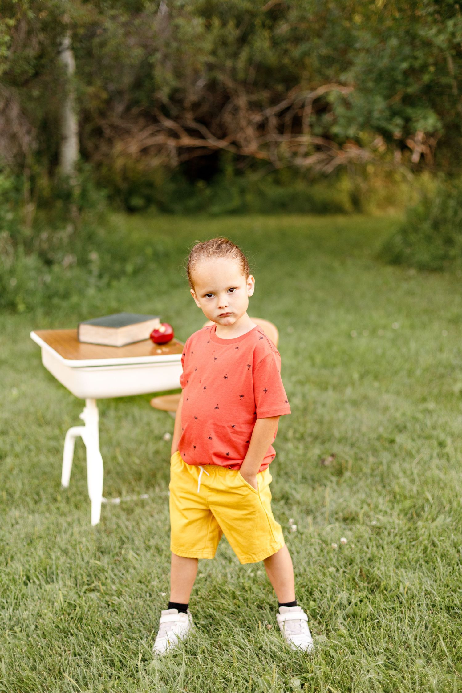 Small child in orange shirt and yellow shorts standing on grass with white side table in background.