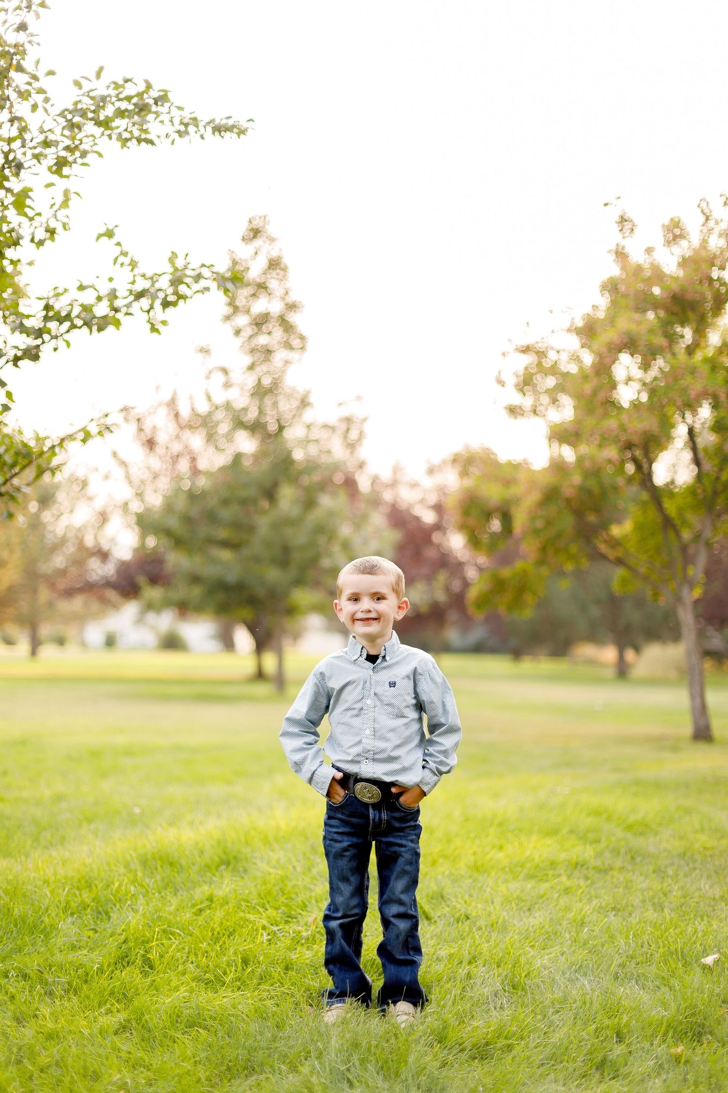 A young child stands in a grassy field wearing blue jeans and a light button-down shirt on an autumn day.