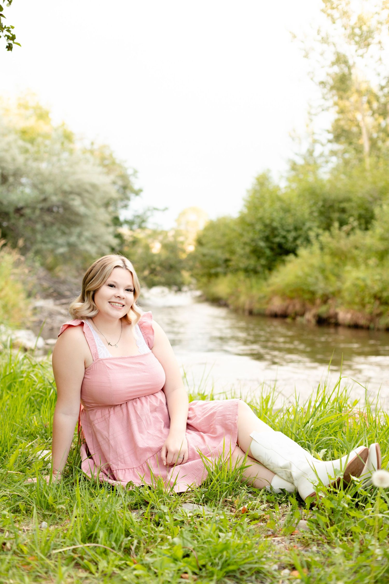 Person in a pink dress relaxes on lush grass by a tranquil stream during a sunny afternoon.
