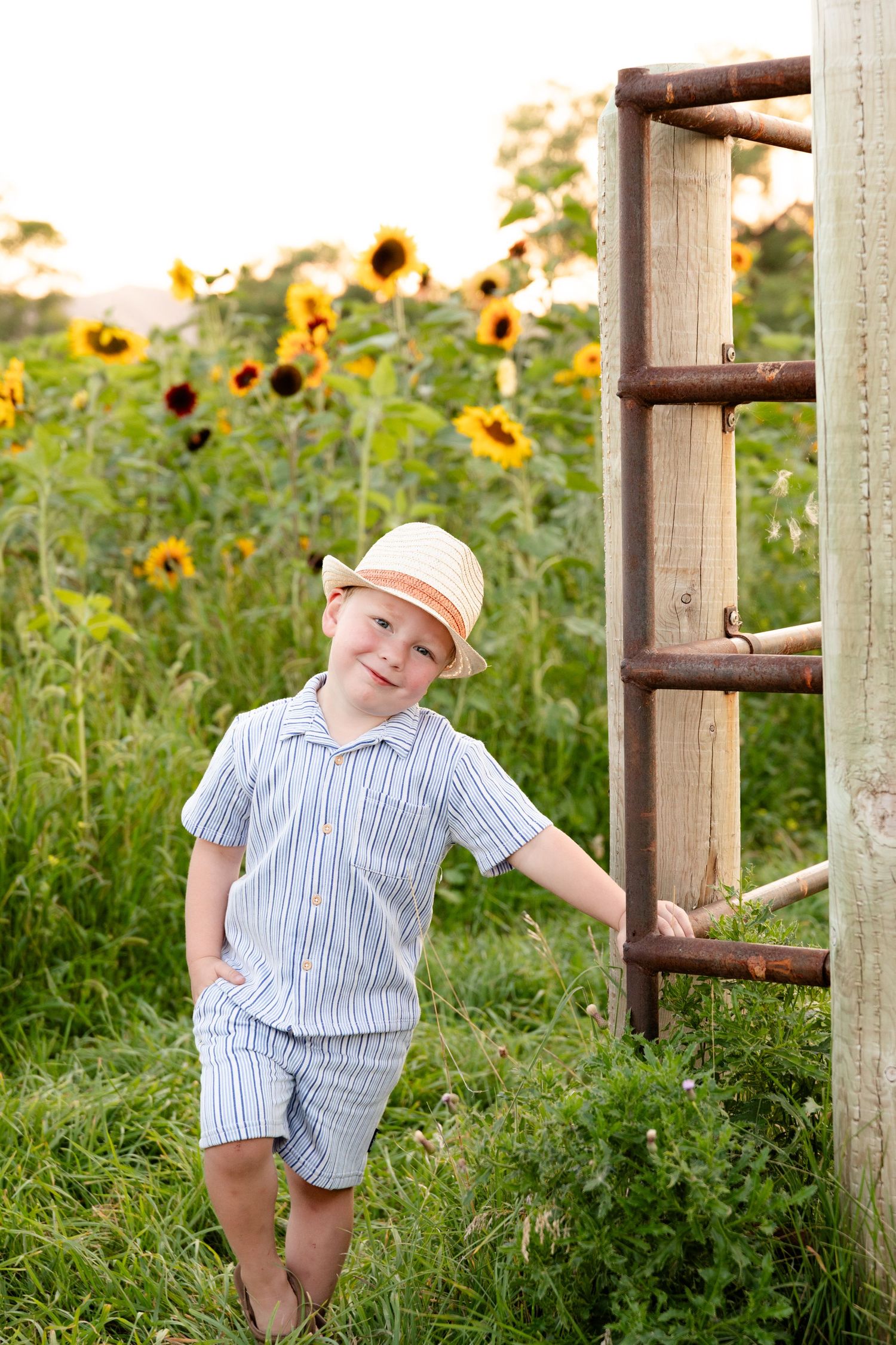 A child in striped romper plays on wooden posts in a sunflower field during golden hour.