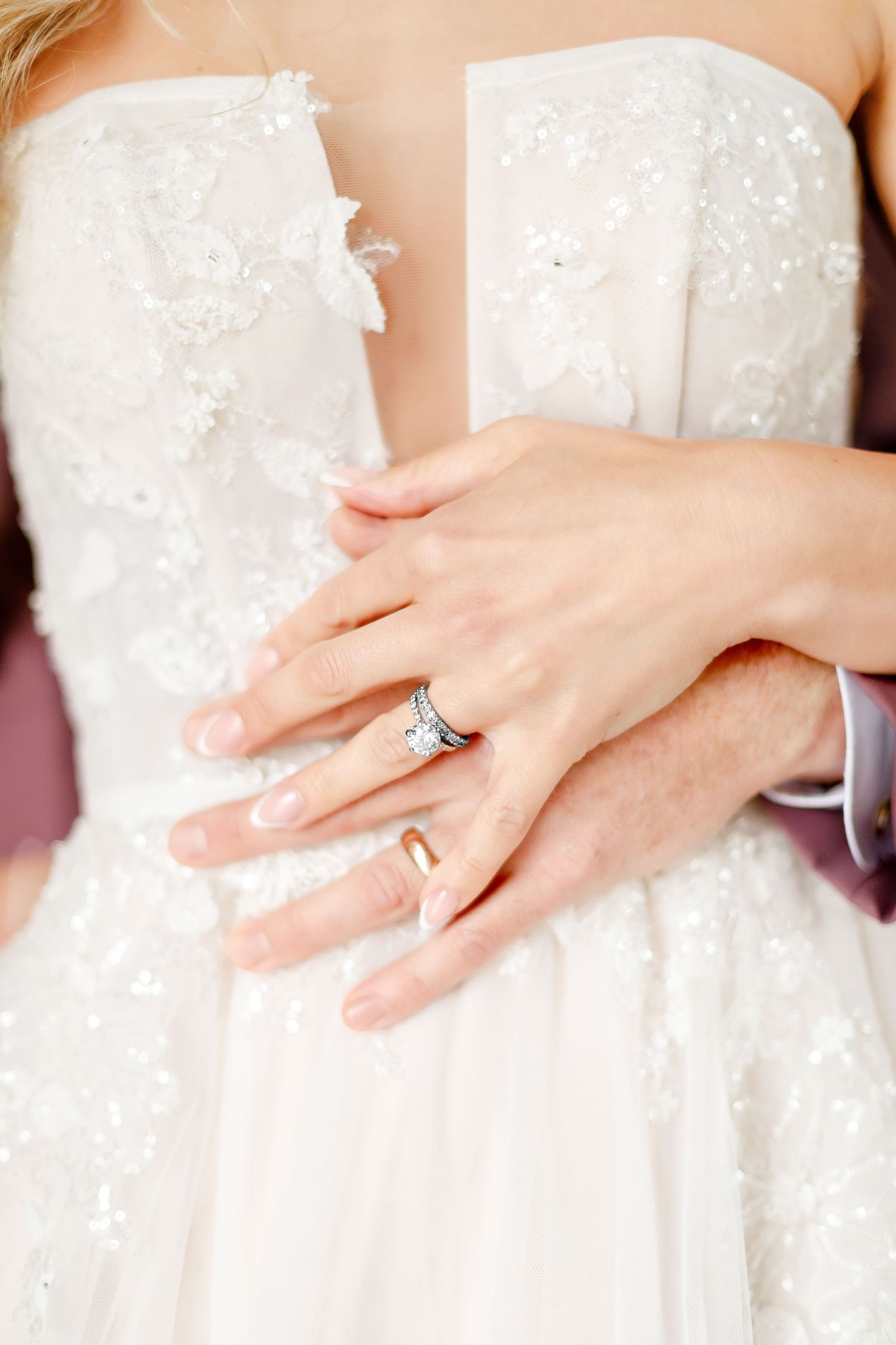 Close up of hands wearing wedding and engagement rings against white lace wedding dress detail.