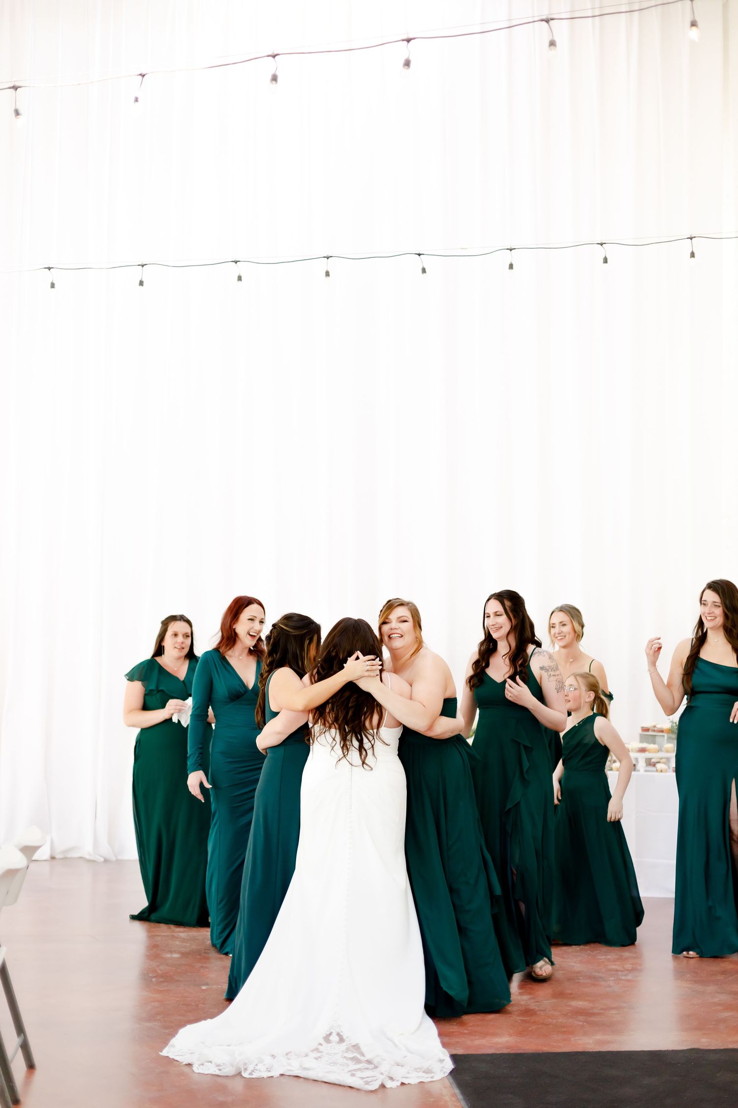 A group of women in emerald green bridesmaid dresses celebrate with bride in white gown in light filled venue.