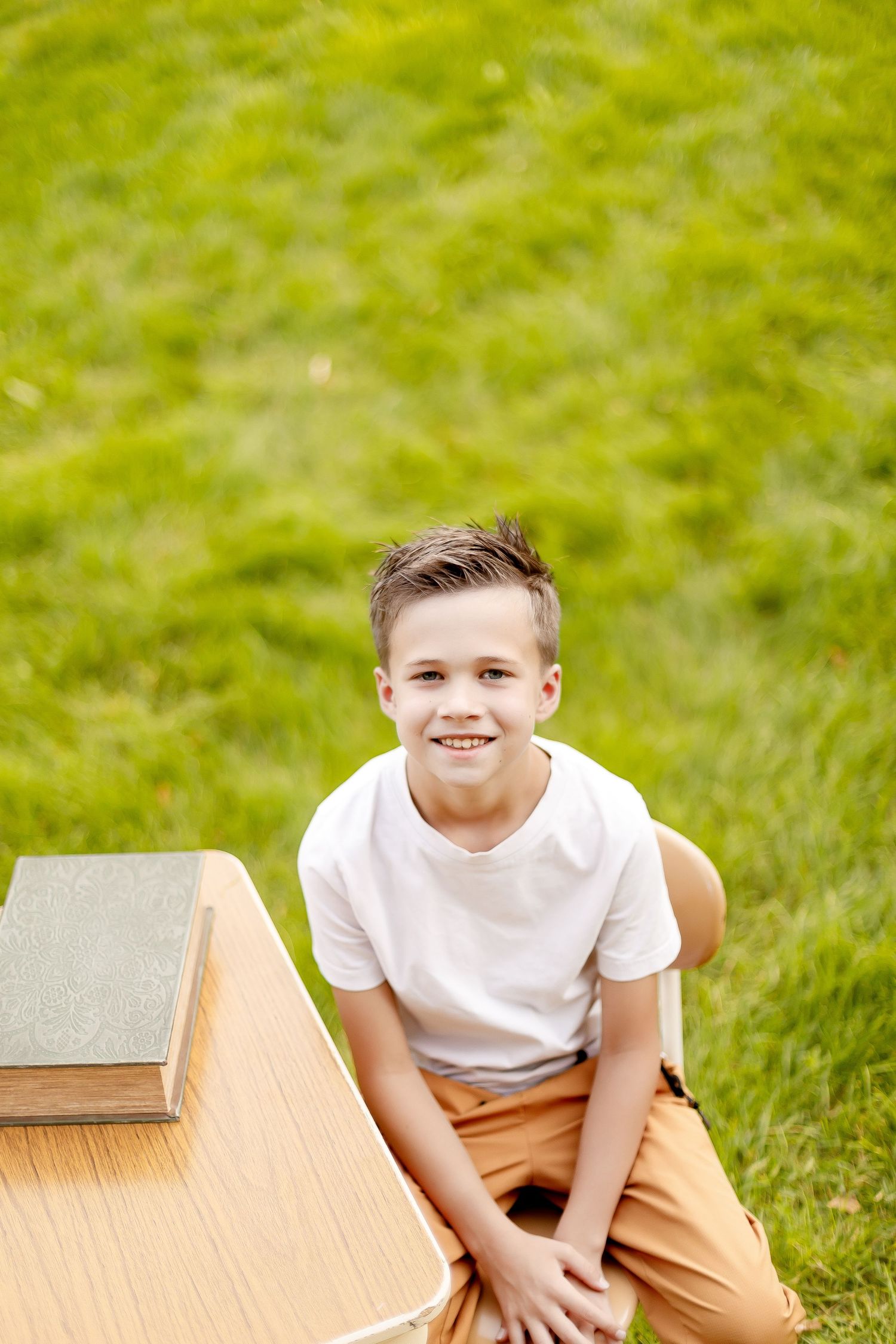 Young student in a white t-shirt sitting on a wooden table outdoors with green grass in the background.