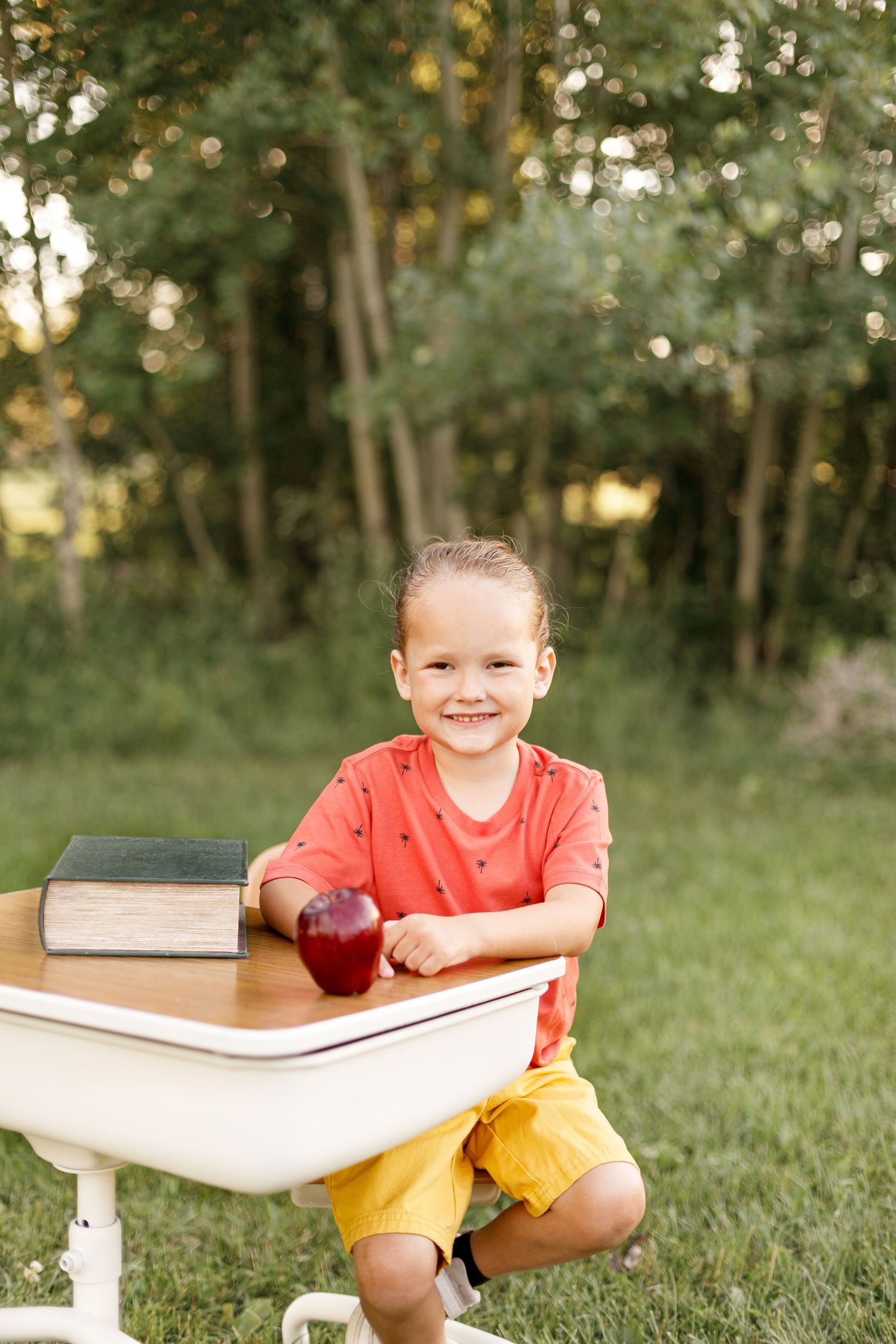A child wearing a coral shirt and yellow shorts poses with books while sitting on white furniture outdoors.