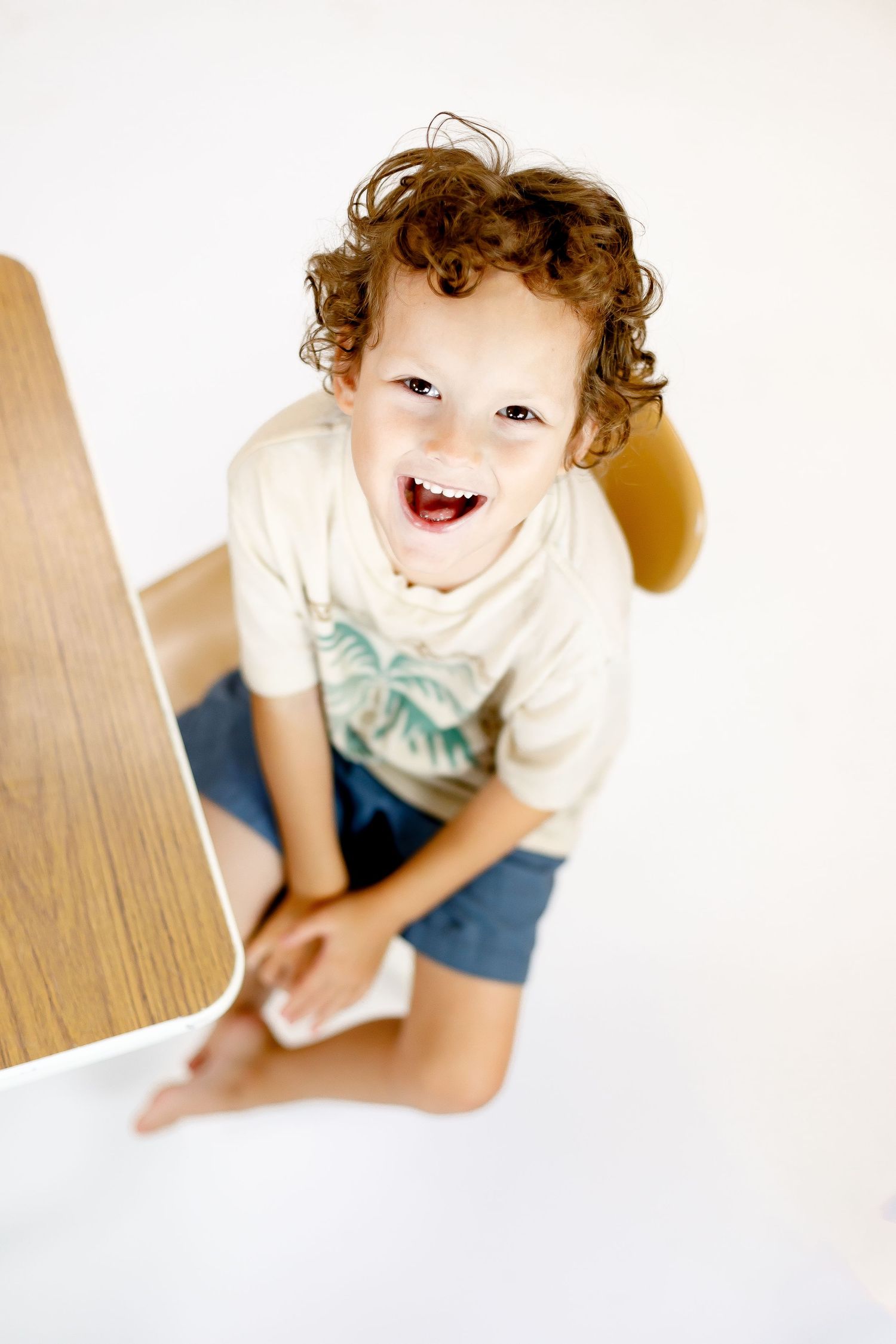 A young child with curly hair laughs while sitting on a wooden floor in a white t-shirt and blue shorts.
