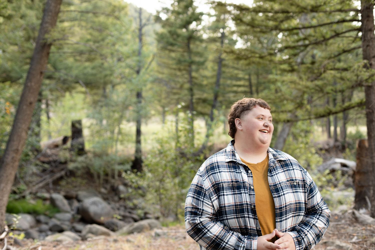Person in a plaid shirt laughs while hiking through a peaceful forest setting surrounded by tall evergreen trees.