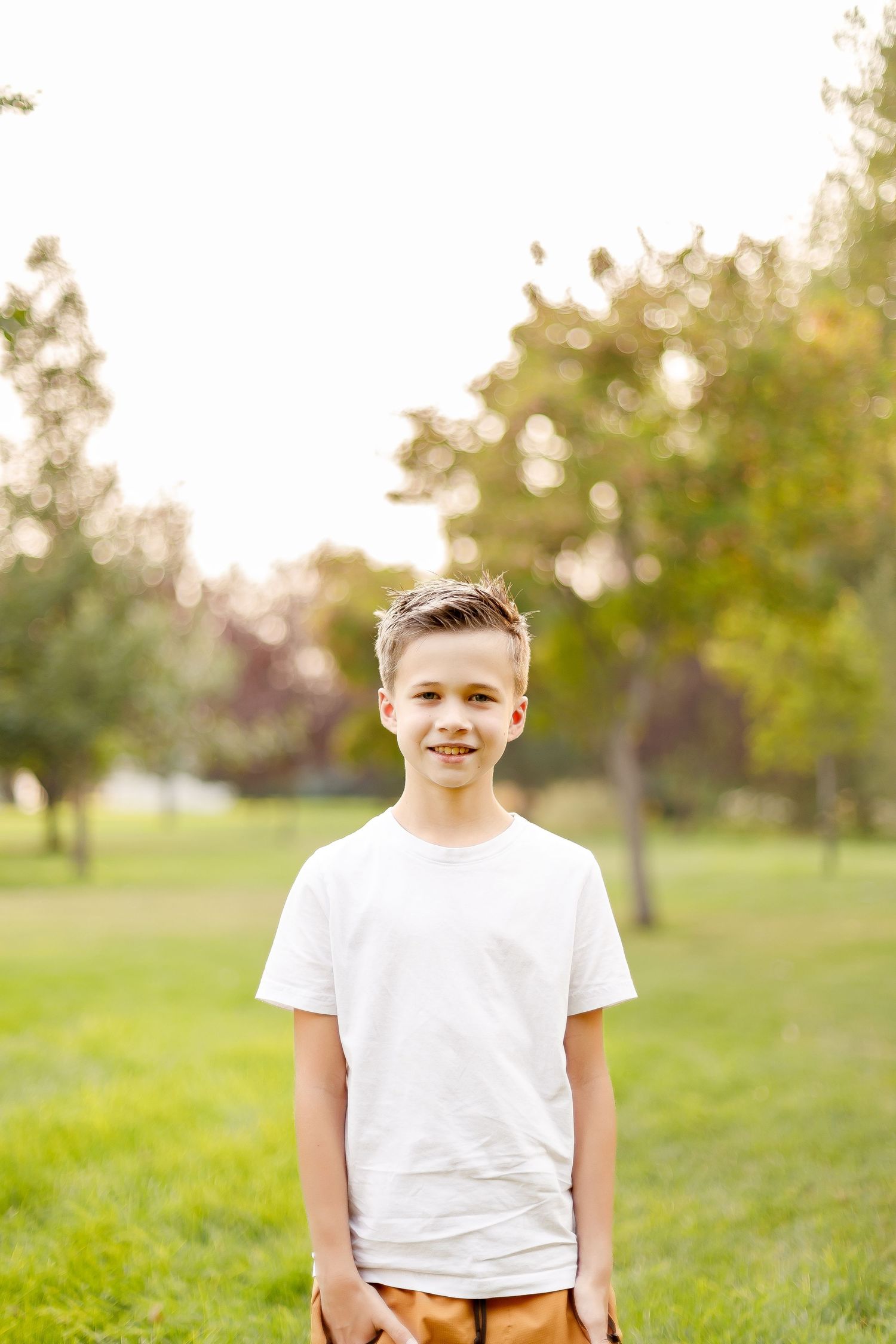 Child wearing a white t-shirt and tan pants stands outdoors in a sunny park with trees in the background.