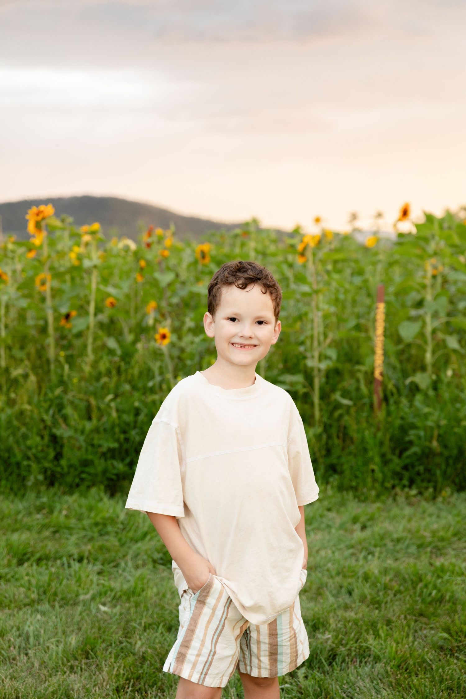 A young child stands in a field of tall sunflowers during golden hour wearing a white shirt and striped shorts.