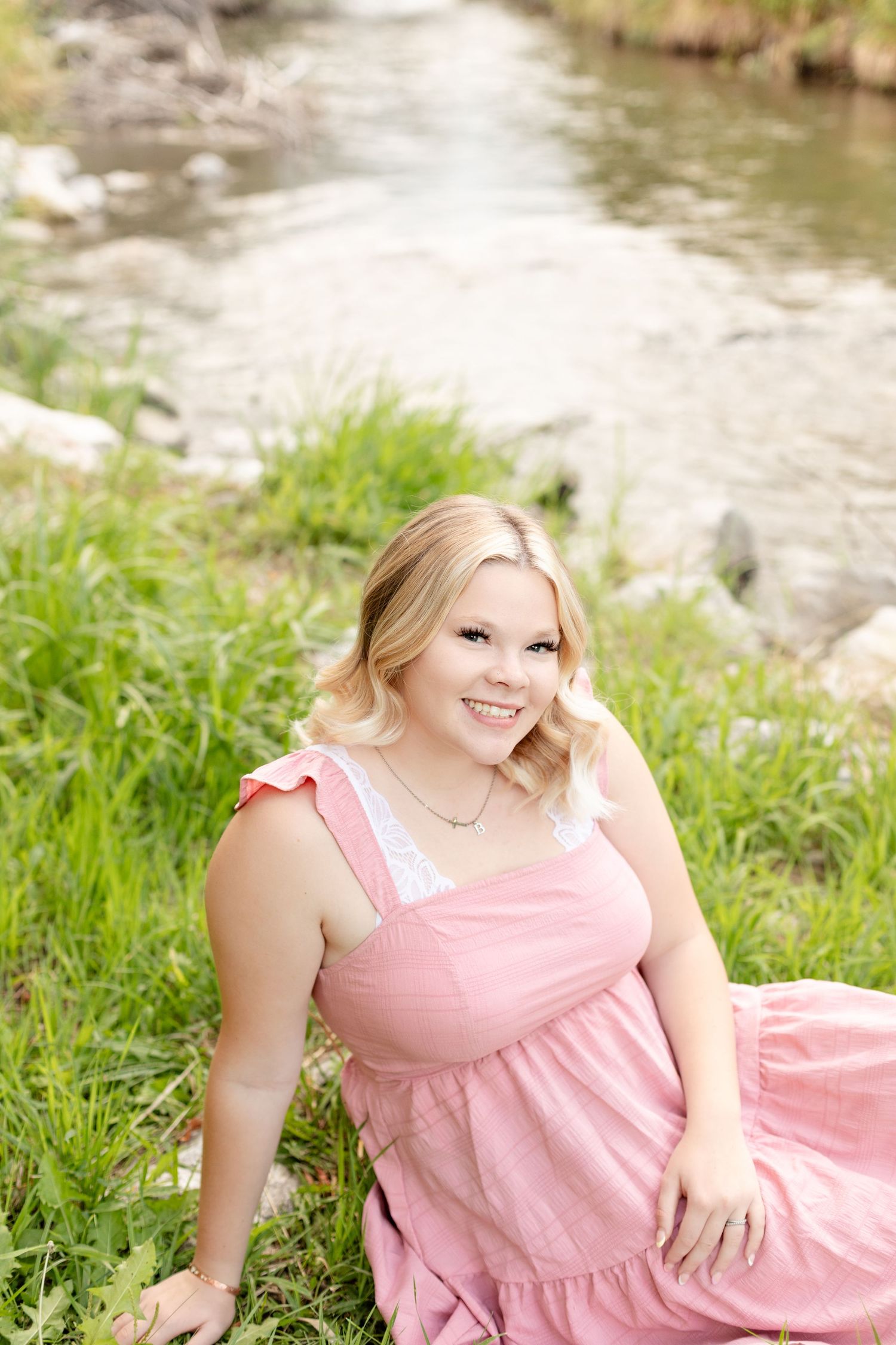 A pink sundress is worn while sitting on grass beside a flowing stream on a sunny summer day.
