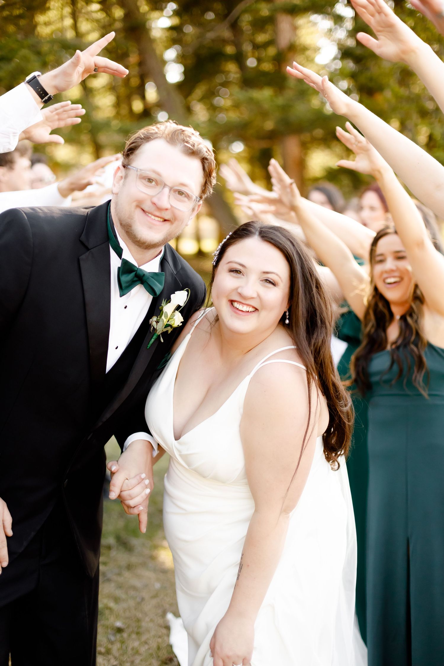 Joyful wedding party celebrates with raised hands as newlyweds laugh together outdoors in natural lighting.