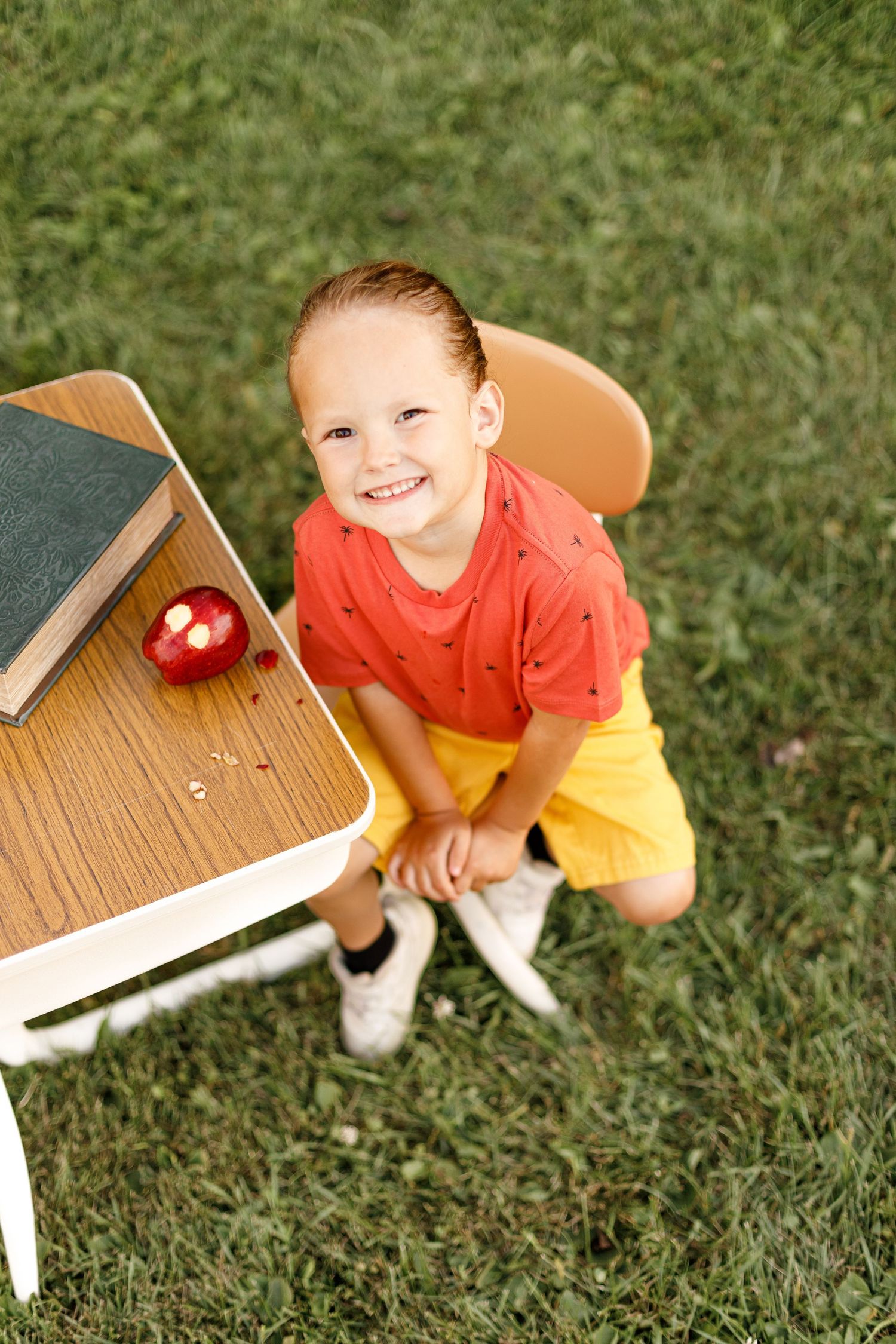 A young child in an orange shirt sits by a white bench in a grassy backyard.