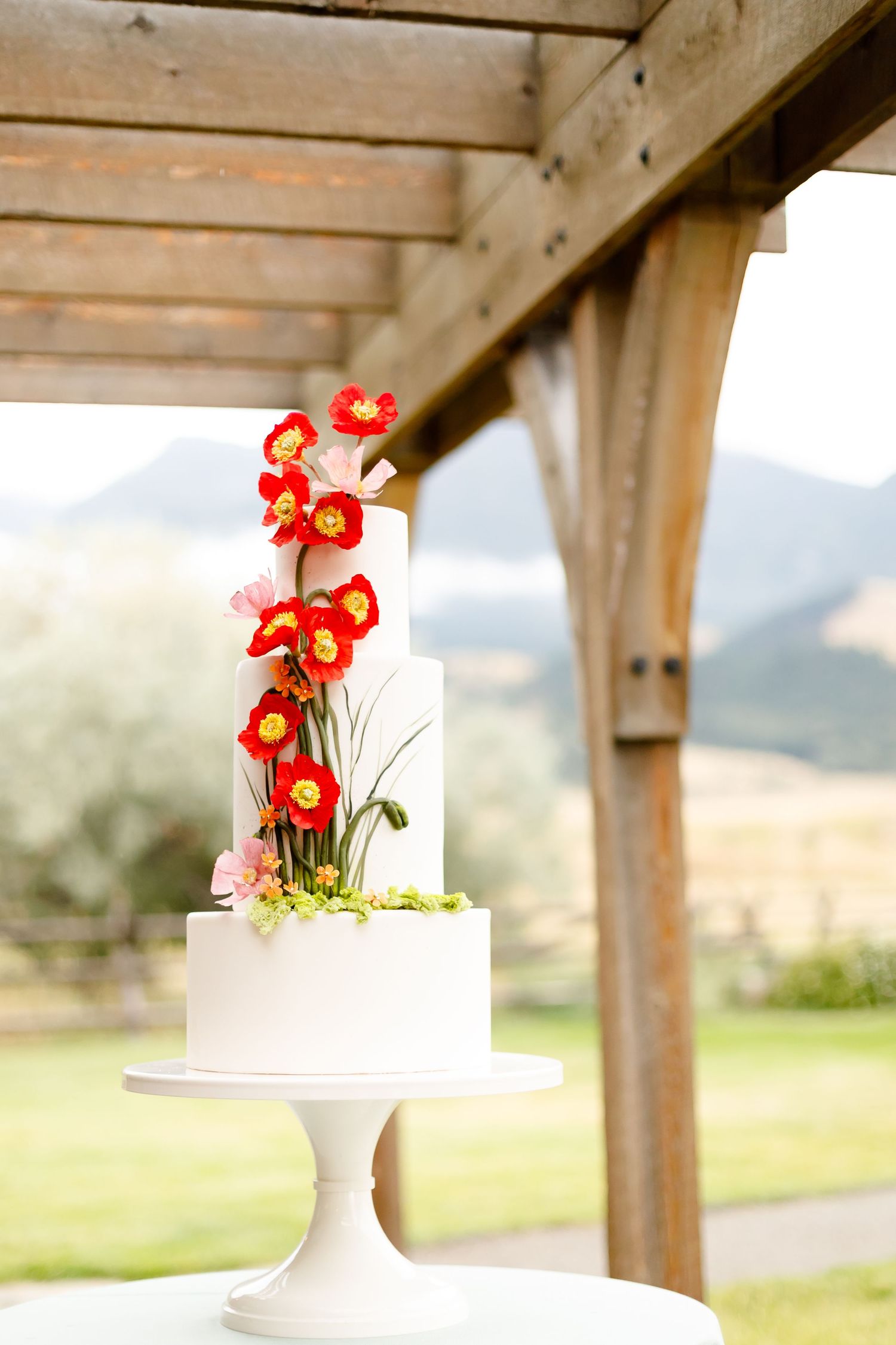 A white wedding cake on a pedestal stand decorated with cascading red-orange orchids under a wooden pergola outdoors.