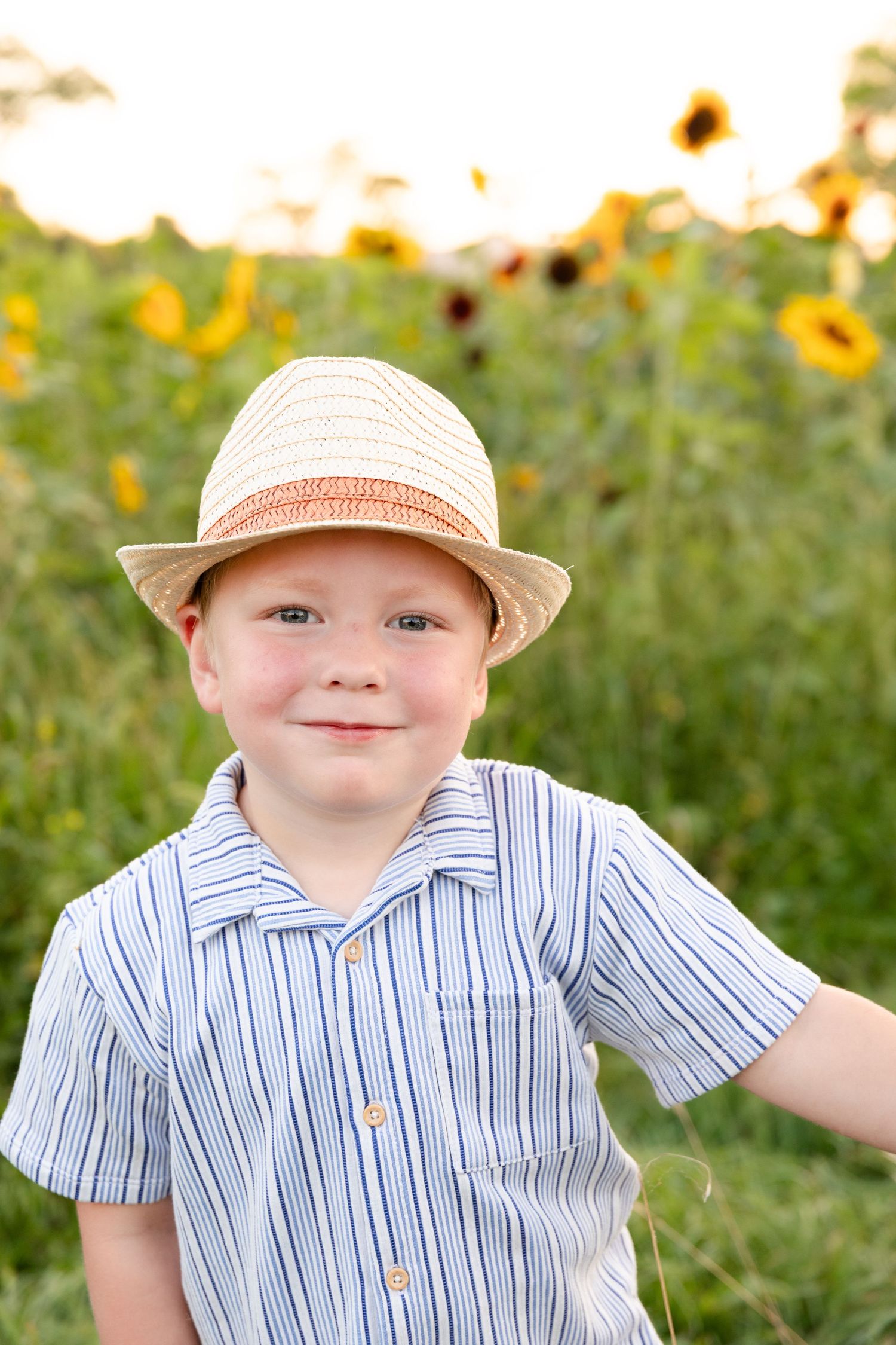 A child wearing a straw hat and striped shirt smiles while standing in a sunflower field on a sunny day.