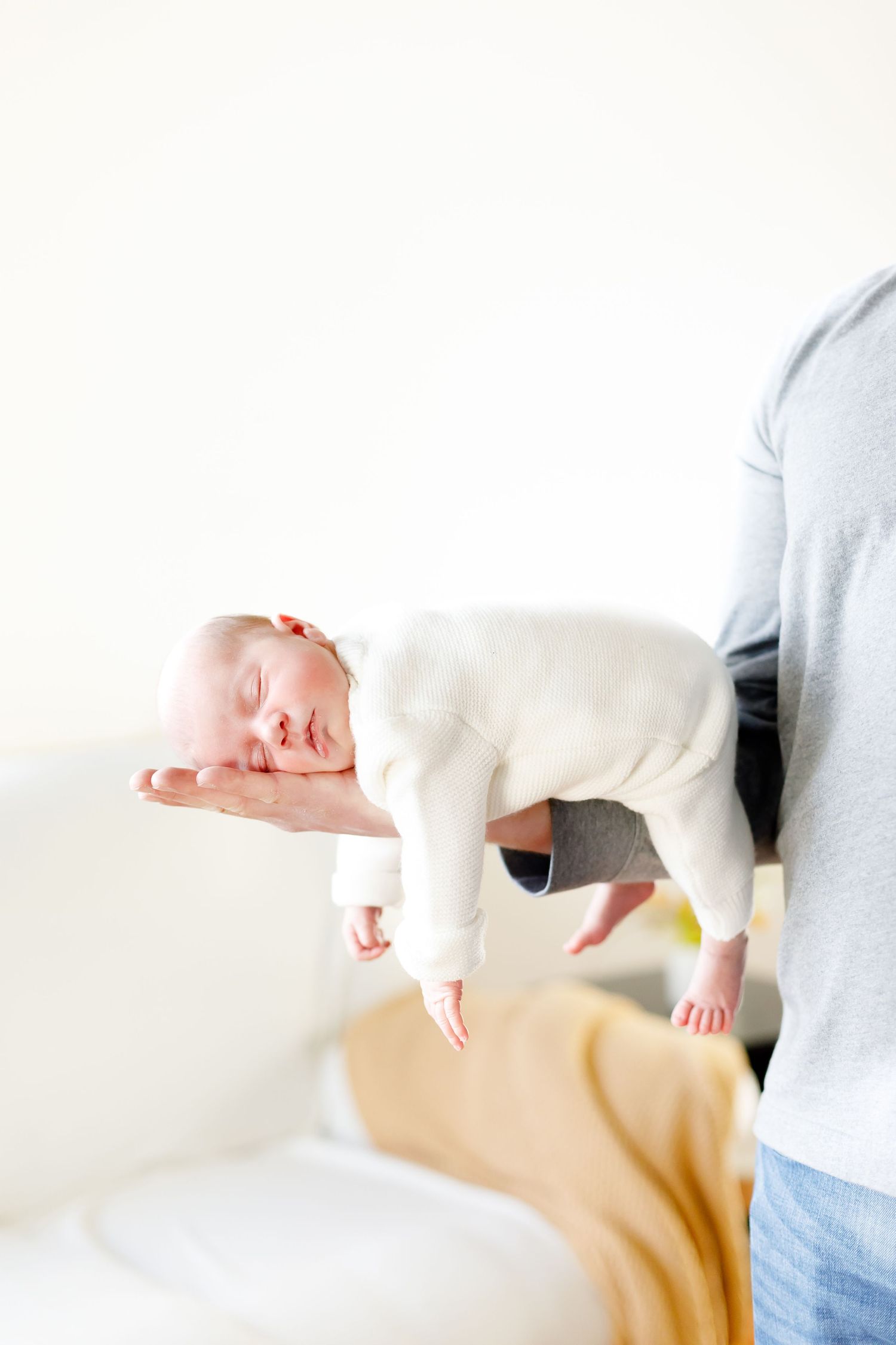 A sleeping baby wearing white clothing is gently held and supported above a bed by adult hands.