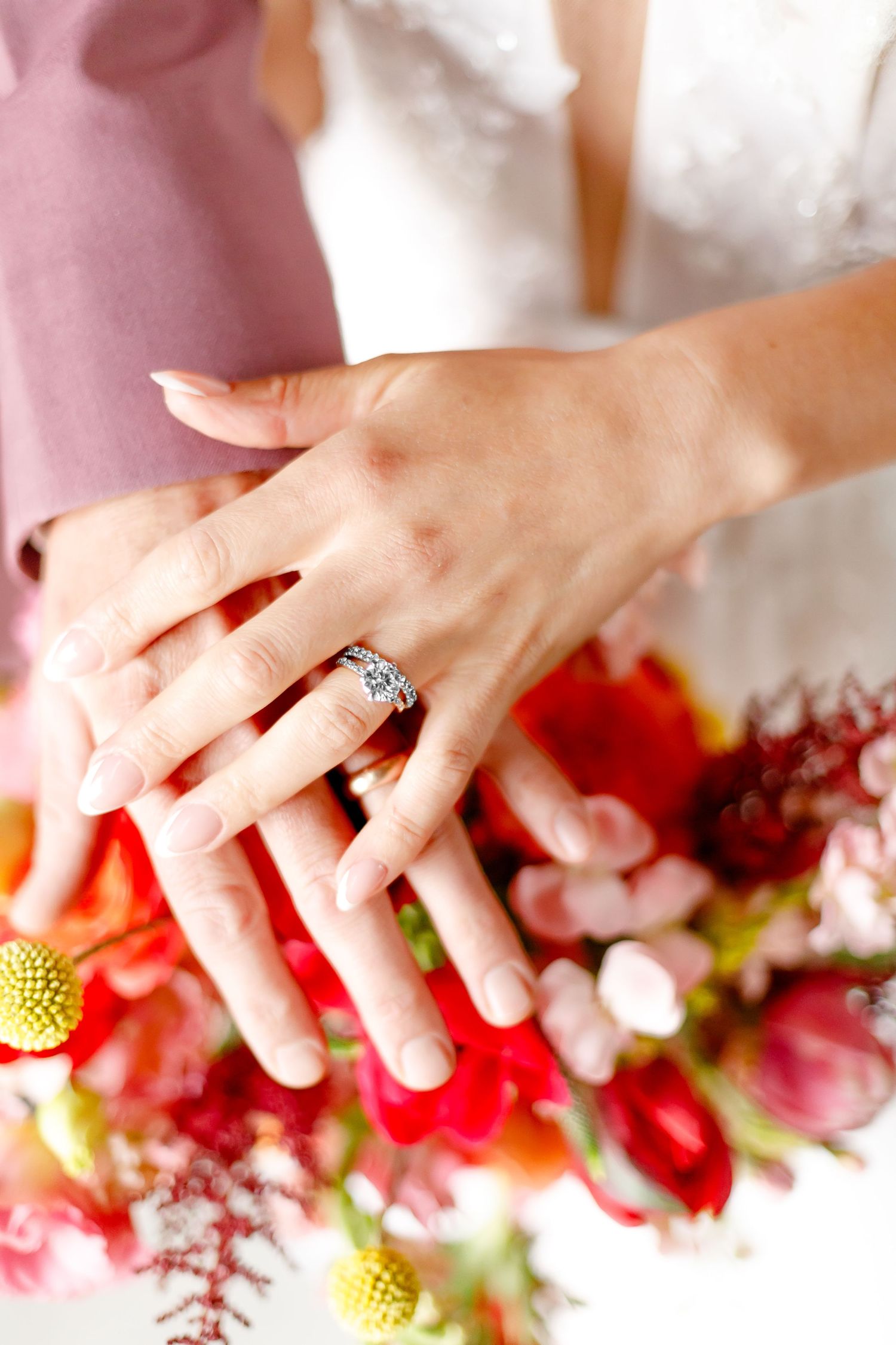 A hand wearing an elegant diamond engagement ring rests against a vibrant bouquet of red and yellow flowers.